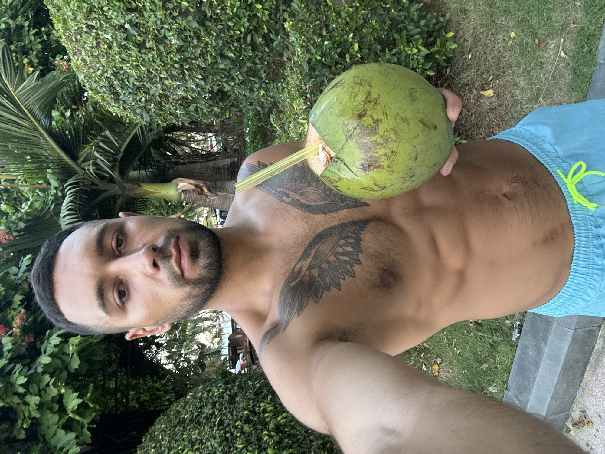 The media file is a selfie taken by a shirtless man in Nha Trang Ward, Vietnam. He is holding a green coconut with a yellow straw inserted into it, appearing to be drinking from it. The man has dark hair, a beard, and visible tattoos on his chest and arm. He is wearing light blue swim trunks.
The background consists of lush green foliage, including palm fronds and bushes, suggesting an outdoor tropical setting. The ground is covered with grass and some fallen leaves. A stone border or pathway is visible to the right. The lighting suggests it is daytime, possibly late afternoon or early evening, with soft natural light. The man's expression is neutral. There is no visible text in the image. The scene depicts a relaxed moment, likely during a vacation or leisure time in a tropical location.