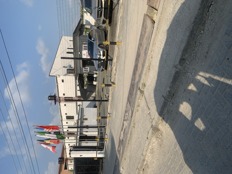 The image depicts an exterior scene in Ikota, Nigeria, focusing on the entrance of a commercial or industrial compound under bright daylight.
In the foreground, a paved road with a concrete curb and an exposed metal drainage cover or ramp runs horizontally. The immediate road surface shows a pattern of concrete pavers, partially covered by dirt, and distinct shadows are cast from the left, indicating the sun's position.
The midground features a perimeter fence constructed of black metal mesh panels supported by black posts, secured by a series of yellow and black striped bollards across the entrance. Behind this barrier, a small, dark grey building, likely a security gatehouse or office, is visible, bearing a sign that reads "THREE ACE GROUP." Two Mercedes-Benz vehicles are parked within the compound, behind the fence: a silver C-Class sedan and a lighter-colored SUV.
The background is dominated by a large, white, multi-story building with a modern, angled roofline. To the left of this building, four flagpoles display the national flags of Canada, Belgium, Nigeria, and the United States, from left to right. A dark water tank on a tall, red-painted metal structure is situated behind the flagpoles. Overhead, power lines traverse the clear blue sky, which features sparse white clouds. No individuals are visible in the scene.