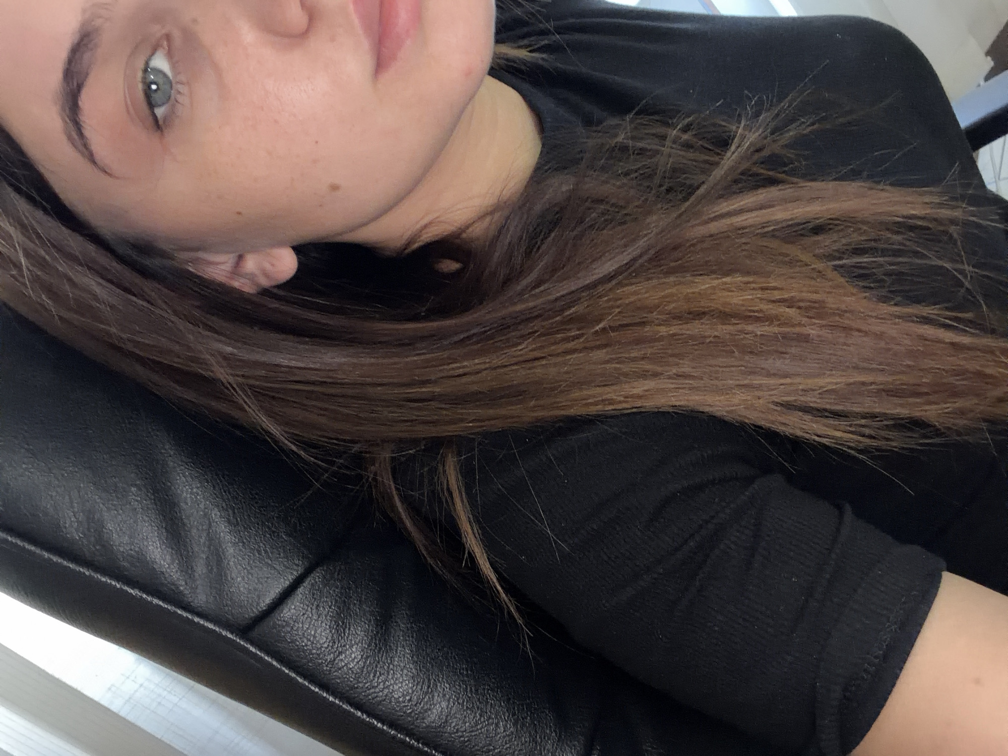 A young woman with long, brown hair, wearing a black t-shirt, is seen from the chest up, with her face turned towards the camera and her head angled to the left. She is seated in a black leather chair, with a portion of the chair visible on the left side of the image. The background consists of a partially visible white blind on the left, and a dark background to the right. The image is a close-up, focusing on the woman's face and upper body. There is no discernible location in Chișinău, Moldova.