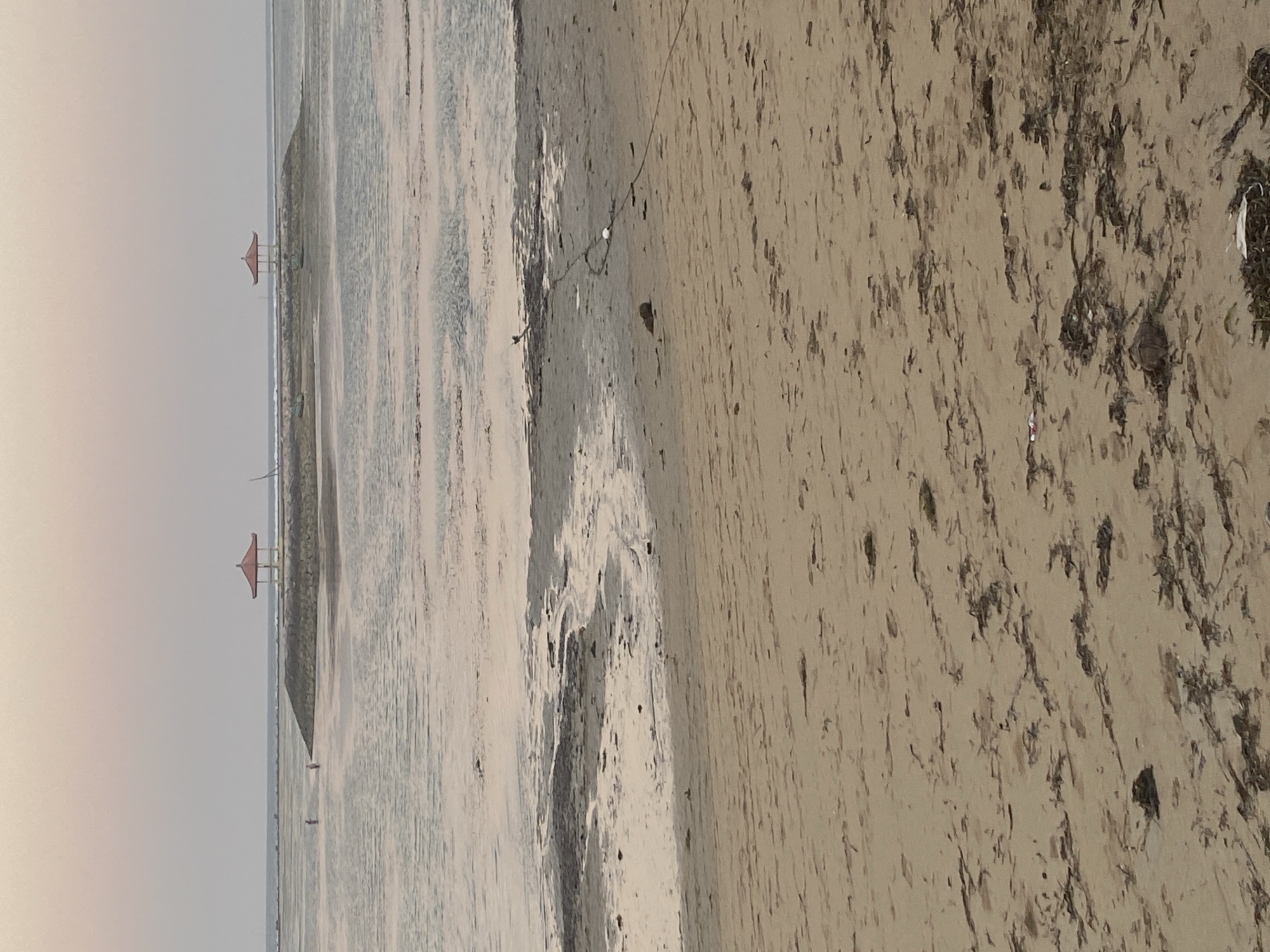 This is a photograph of a beach in Denpasar, Indonesia, likely taken during low tide. In the foreground is a sandy beach with scattered seaweed and debris. In the middle ground, the shallow water reflects the sky and the seabed is visible in patches. Further out, two gazebos with red roofs stand on a stone structure extending into the water. Two figures can be seen walking in the shallow water near the stone structure. The sky is a pale pinkish-grey, suggesting it is either dawn or dusk.