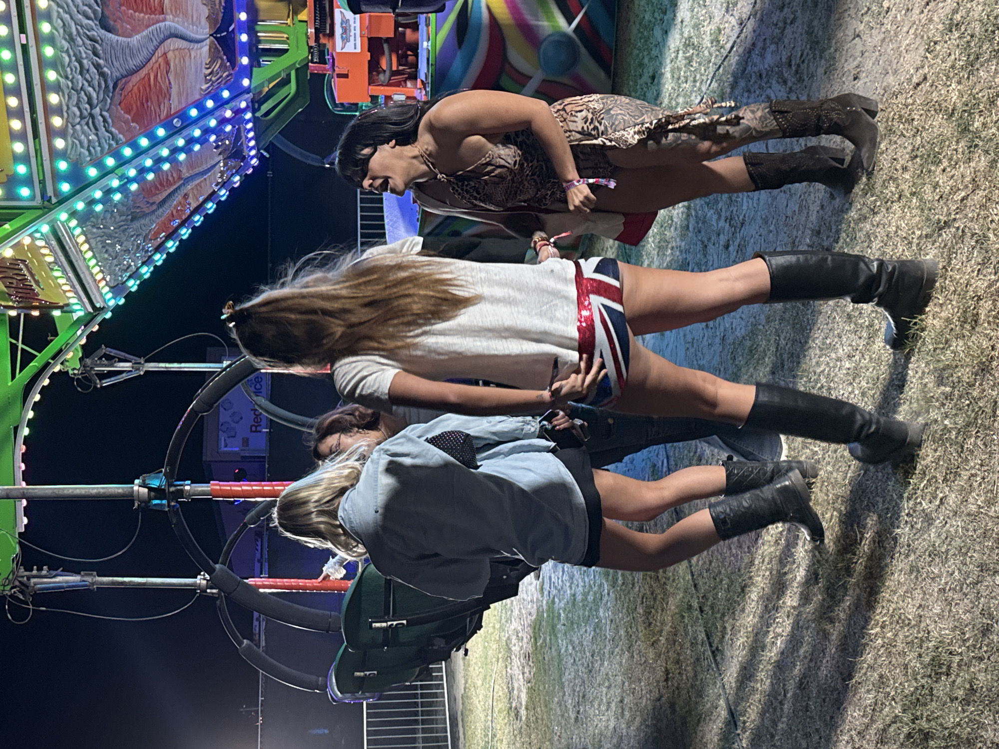 The image shows three women standing on a grassy area at night. They appear to be at an outdoor event or fair in Thermal, United States of America. The woman nearest the camera is wearing a denim jacket, a black skirt, and black boots. The woman in the middle is wearing a white shirt and shorts with the Union Jack flag on the back, paired with black boots. The woman farthest from the camera is wearing a leopard-print dress and tan boots. In the background is a carnival ride with colorful lights.