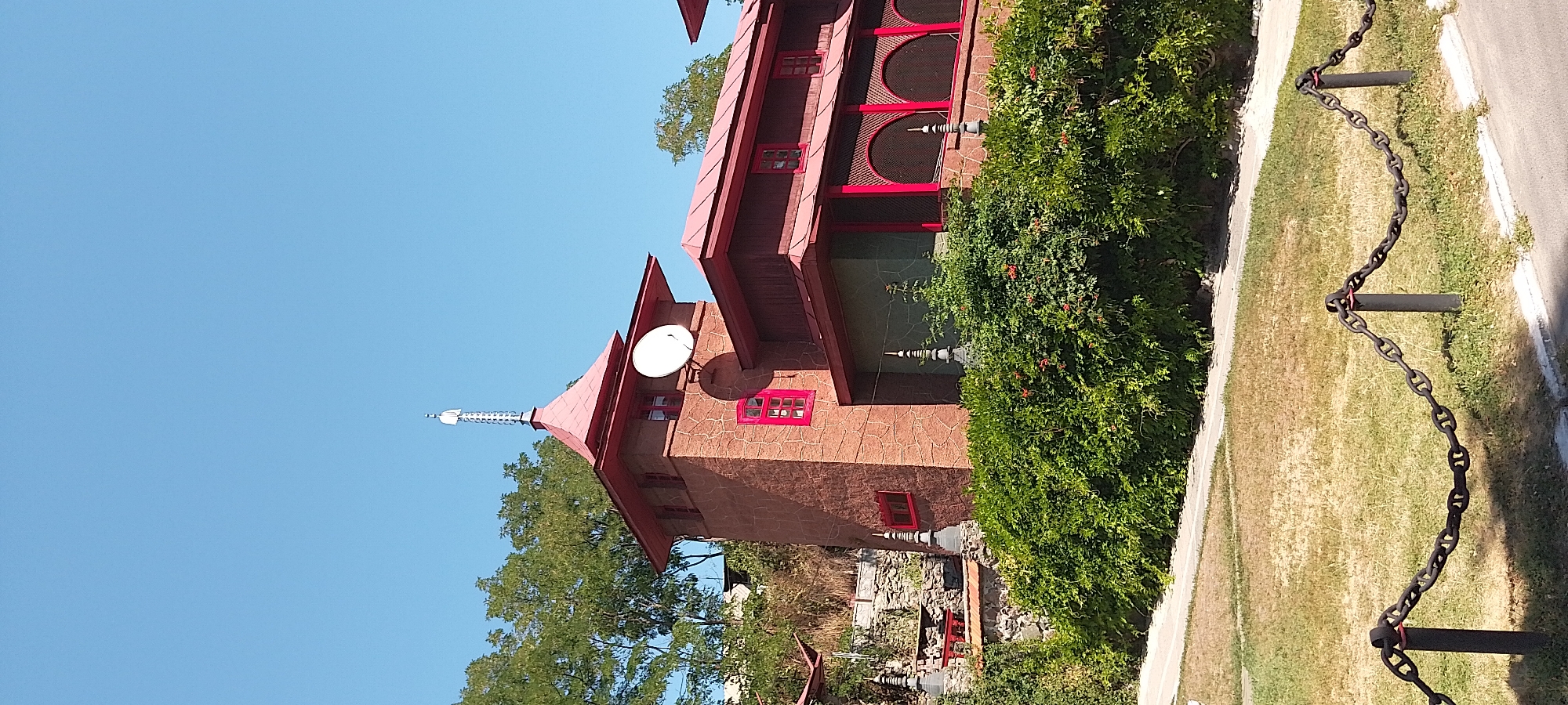 The image shows a distinctive building with a red roof and brown brick facade, featuring multiple levels and a tower topped with a spire-like antenna. A satellite dish is mounted on the side of the tower. The building is surrounded by lush green bushes and some dry grass in the foreground, with a decorative chain fence in the lower part of the frame. The sky is clear and blue. This building is located in Cherkasy, Ukraine, and is identifiable as the Cherkasy Regional Museum of Local Lore.