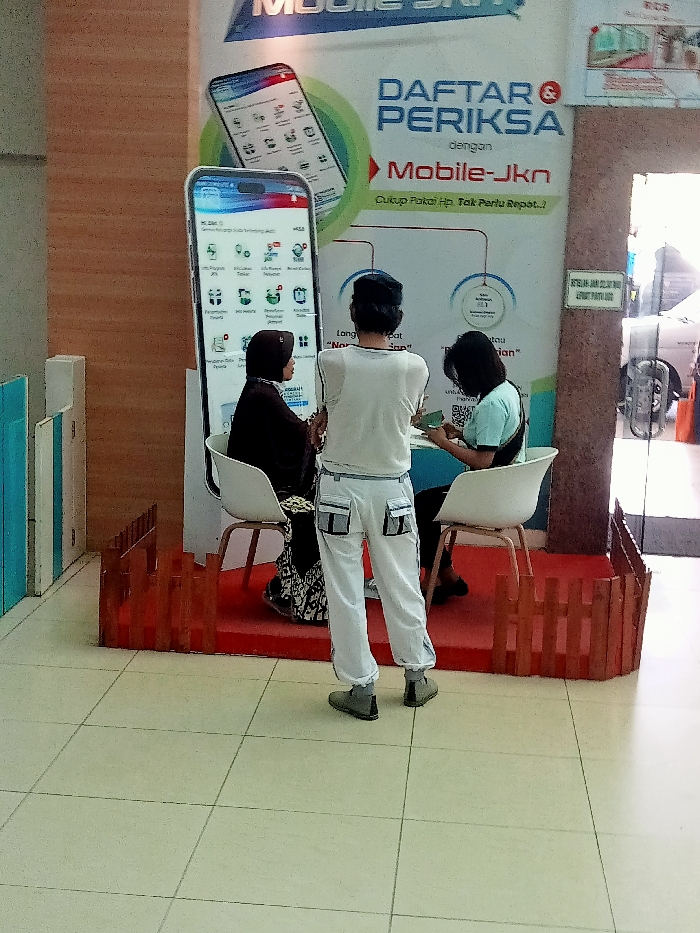 The image shows a scene inside a building in the Kecamatan Margorejo district of Surabaya, Indonesia (latitude: -6.76, longitude: 111.015). Three people are sitting at a table in front of a large advertisement. The ad is for a service called "Mobile-Jkn" and is decorated with a large digital phone display showing a list of icons. The phone is positioned to the left of the table. The person in the center is a man wearing a white shirt and white pants. He is facing the camera and looks to be talking to the woman sitting to his right. The woman has short, dark hair and is wearing a blue shirt. She is looking down at the table, presumably at a document. The woman seated to the left of the man is wearing a black headscarf and black dress. She is looking towards the phone. All three people appear to be interacting in some form of transaction. The scene is set on a raised platform, which has a wooden fence along the front. The floor is tiled, and the walls of the room appear to be a light color. There is an open doorway to the right of the scene which allows a glimpse into another room. The overall scene suggests an office or other business setting.