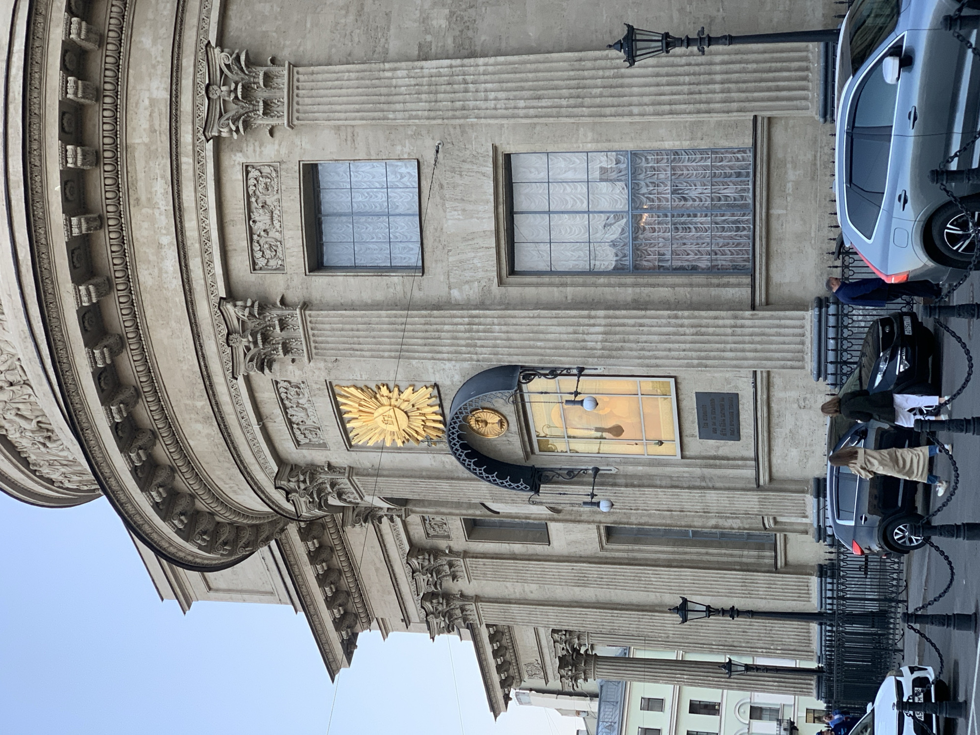 The image shows a partial view of the architectural facade of the Saint Isaac's Cathedral in Saint Petersburg, Russia. The building exhibits neoclassical design elements, including detailed cornices, fluted columns, and ornate decorations. Two window openings are visible on the facade, as well as a gilded sun-shaped emblem and a decorative canopy above an entrance. Pedestrians are present near parked vehicles along the building's perimeter. A lamppost and a decorative fence line the foreground. The sky is a pale blue, suggesting clear weather.