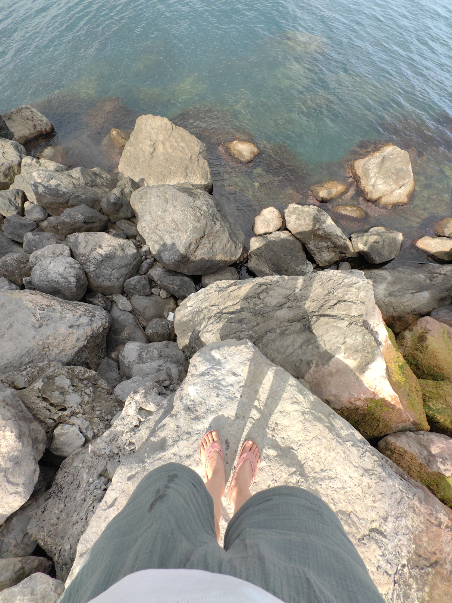 The image shows a first-person perspective of a person standing on large rocks near the sea. The person is wearing green pants and pink flip-flops. Their feet are positioned on a large, light-colored rock, and their legs are visible from the knees down. The surrounding area is filled with numerous gray and beige rocks of varying sizes, some partially submerged in the clear, blue-green water. The water appears calm, with gentle ripples on the surface. Sunlight casts shadows from unseen objects onto the rocks, suggesting it is daytime. The presence of rocks and the sea indicate a coastal environment, likely a rocky shore or a breakwater. There are no visible signs of text or any specific activities other than standing in the location. The scene conveys a sense of peace and natural beauty.