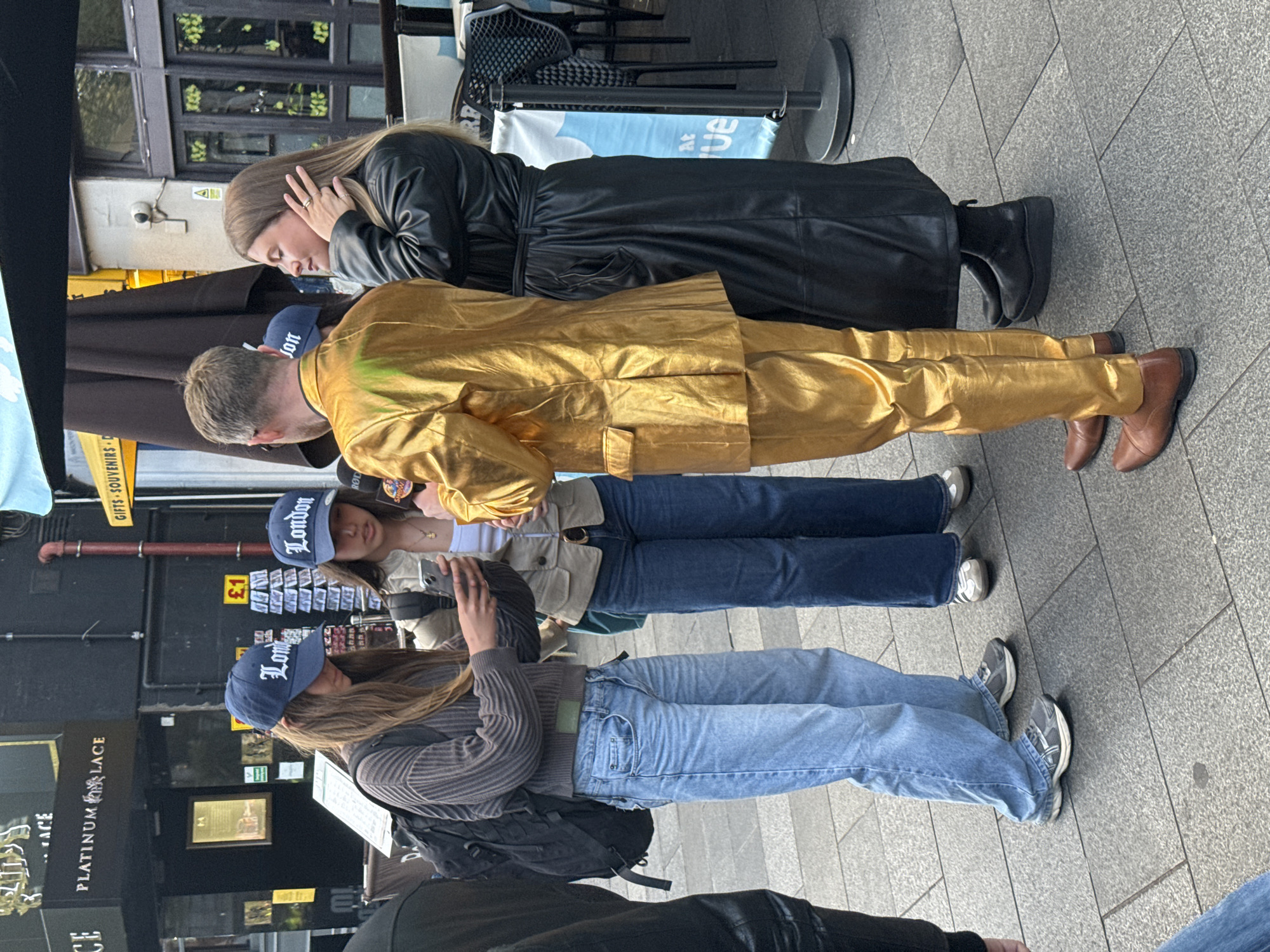 The image captures a group of people standing on a tiled city pavement near a store, likely in London, United Kingdom, based on the signage and style. Four people are predominantly featured: one in a black leather coat, another in a golden suit, and two others wearing blue baseball caps with the word "London" embroidered on them. The two people wearing the hats are also wearing jeans. One of them is also wearing a grey sweater, and the other is wearing a white top and beige jacket. The group appears to be engaged in conversation or observation. Store signage includes "PLATINUM LACE" and "GIFTS SOUVENIRS." Other details visible are a trash can and some chairs.