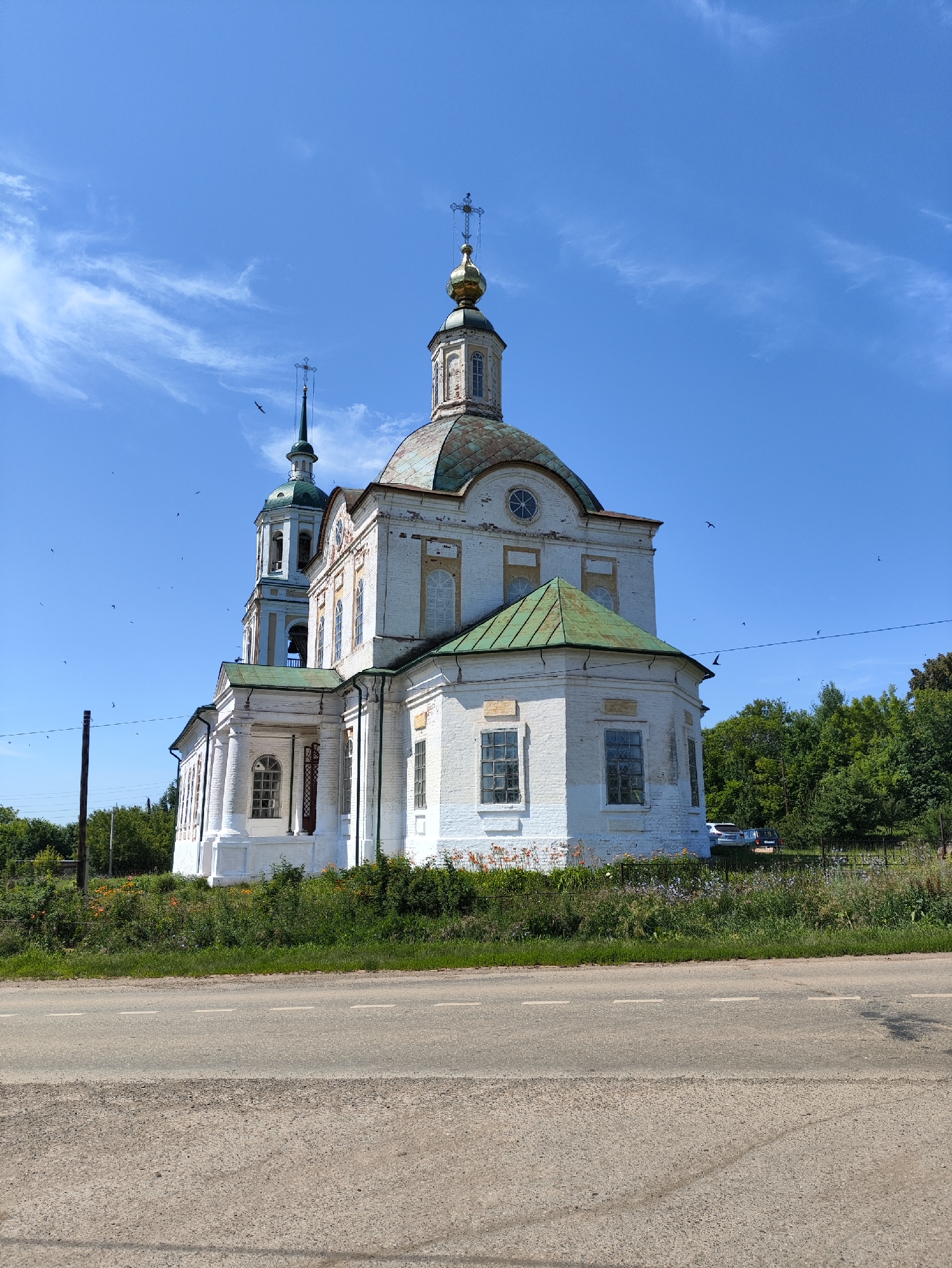 A white brick church with a green roof and a golden dome stands against a clear blue sky dotted with wispy clouds and several birds in flight. The church has a bell tower with a green spire and a cross on top, and another dome with a cross on top of the main structure. The building is adorned with arched windows and decorative elements. In front of the church, there is a patch of green grass and flowers. A road runs along the bottom of the image, with a granular texture. In the background, a dense line of green trees forms the backdrop, and two cars are visible parked near the trees. The overall scene is a rural landscape bathed in bright daylight, suggesting a pleasant, clear weather day.