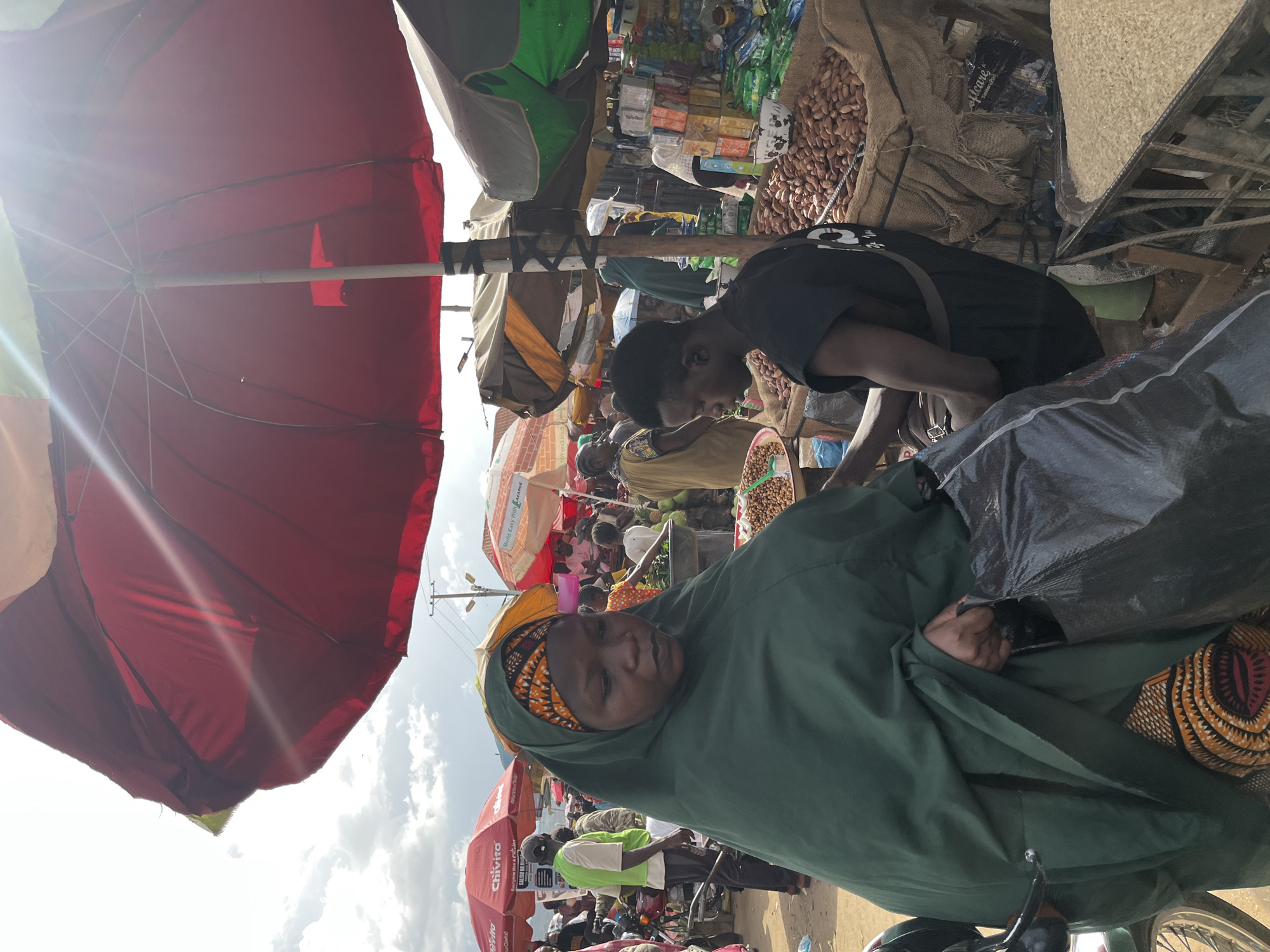 The image depicts a bustling outdoor market scene, likely in Rafin Par, Nigeria. A woman wearing a dark green hijab and an orange and black patterned headscarf is in the foreground, facing left. She is holding a large black plastic bag in her right hand. To her right, a young man in a black t-shirt is partially visible, seated and appearing to be tending to goods. He has a strap across his chest. In front of him are large sacks filled with what appear to be nuts or seeds, and a metal grate holding a large amount of rice. Behind the woman, a variety of market stalls and people are visible under large, colorful umbrellas, including red, yellow, and green ones. Some stalls display packaged goods. The overall environment suggests a lively and active marketplace.