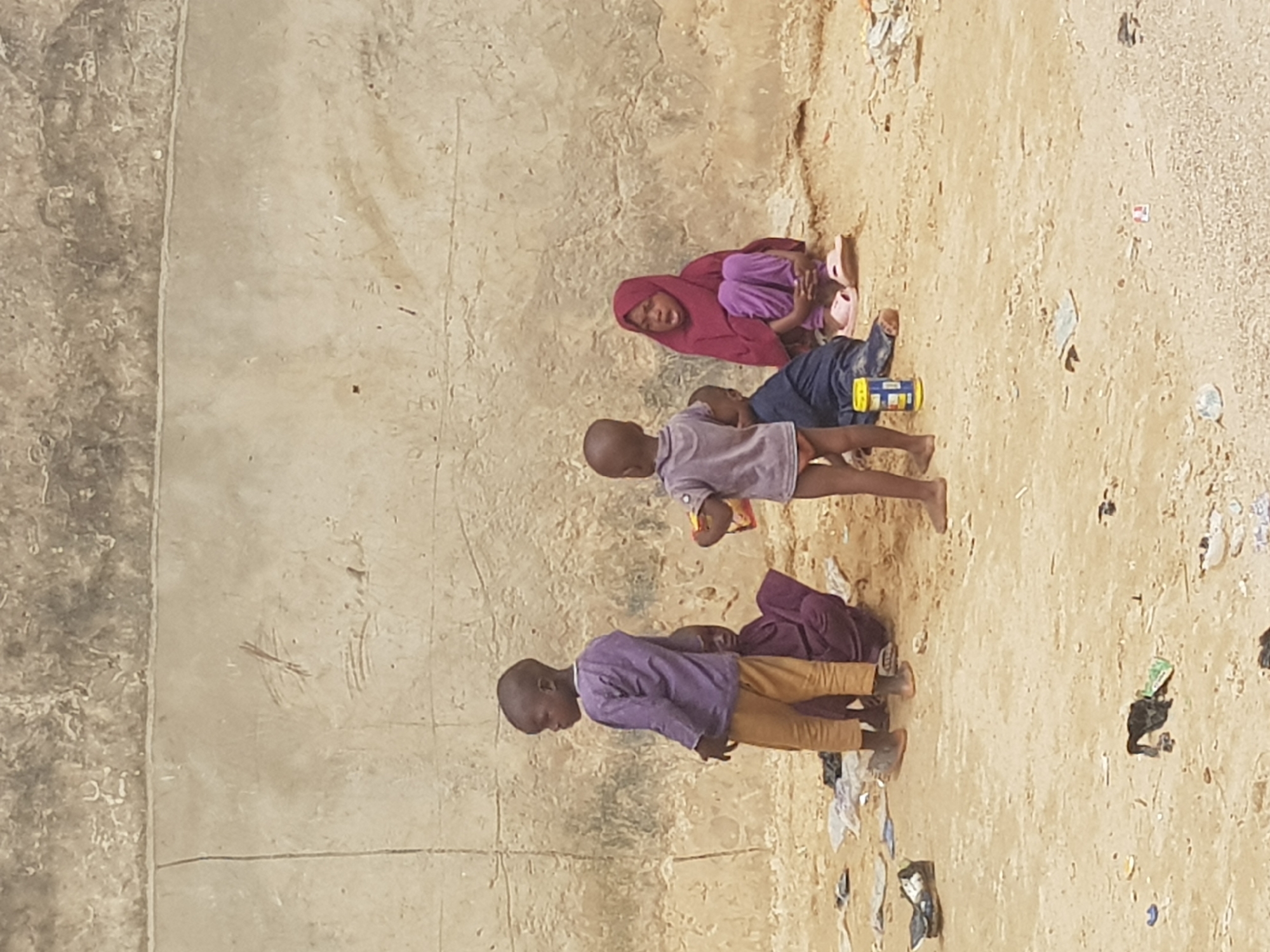 Four children are on the ground in Dutsinma A, Nigeria. The ground has a concrete section and a sandy, littered section. One child wears a maroon head covering, lying with another wearing a purple shirt and blue pants. Another child wears a purple shirt, lying on their stomach. The fourth child wears a purple shirt and tan pants and is standing.