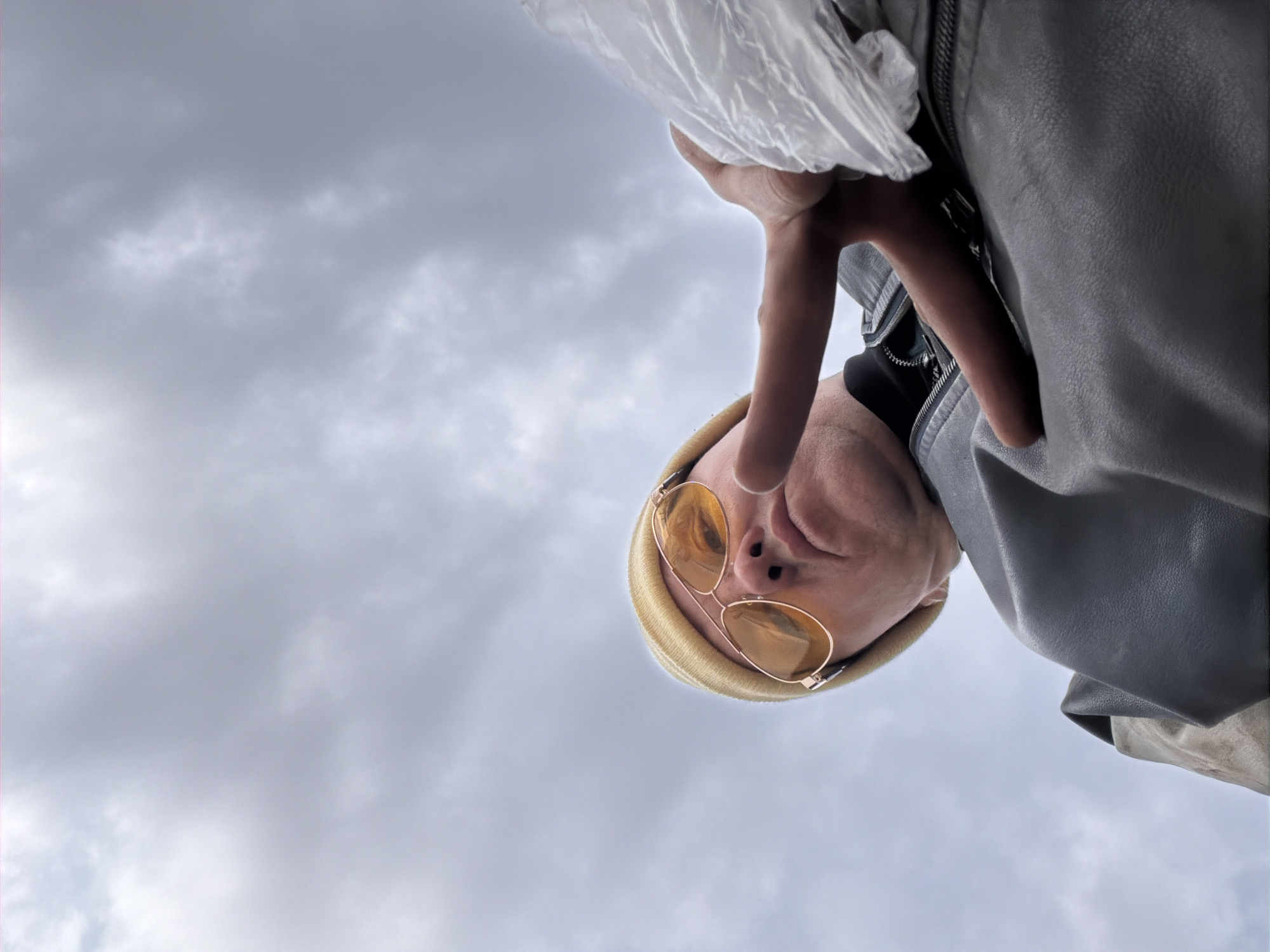 A low-angle photograph depicts a person wearing gold-rimmed sunglasses and a tan beanie. The individual is partially framed by a grey leather jacket. A hand holding a translucent plastic bag extends into the frame. The background features an overcast sky. The city of Chaikovsky, Russia is not discernable from the image.