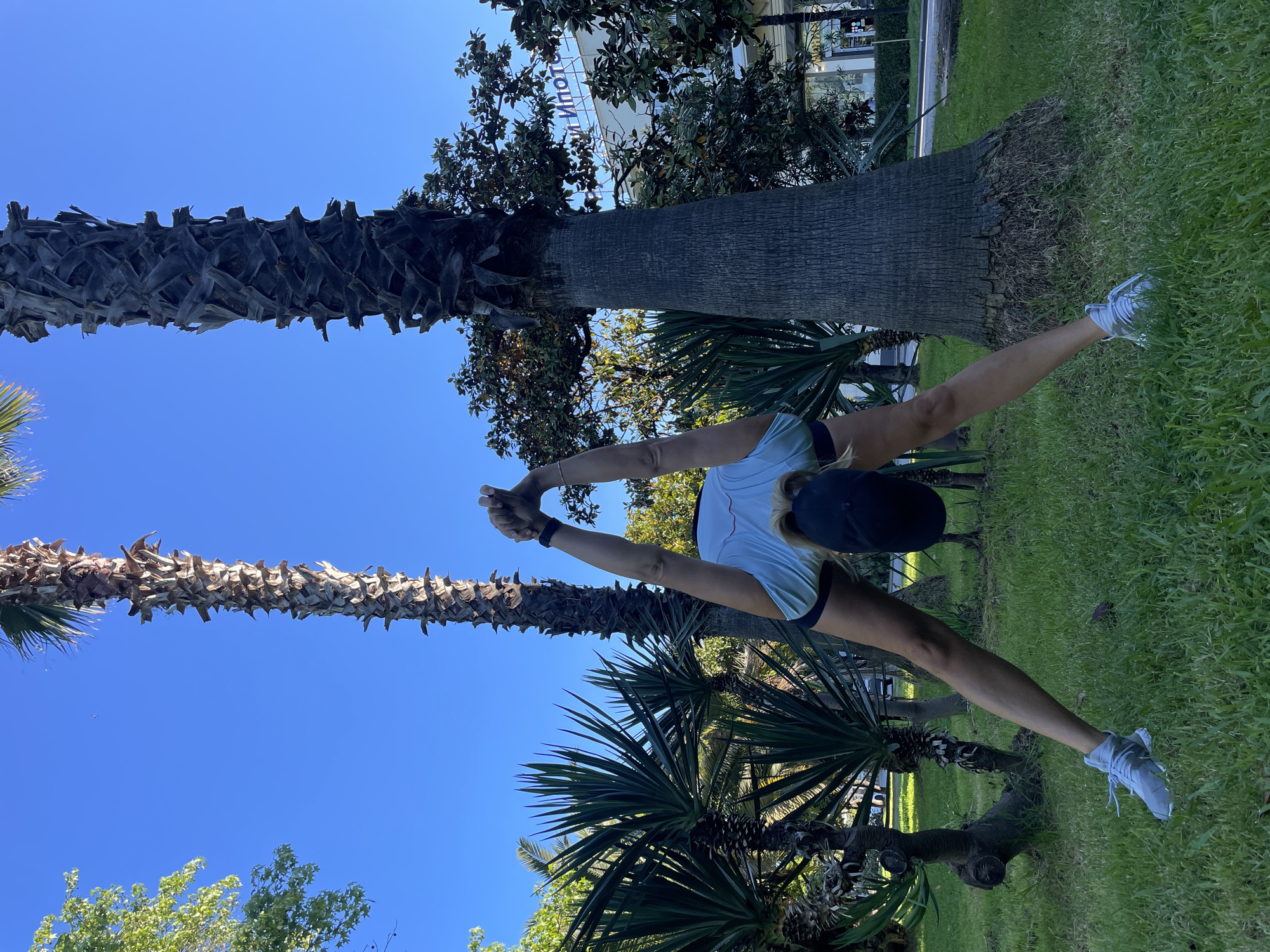 The image displays an outdoor scene featuring a person performing a physical stretch under a clear blue sky. The central subject is a person, likely female, dressed in light athletic wear including a light-colored top, dark shorts, a dark baseball cap, and light-colored athletic shoes. A fitness tracker is visible on their left wrist. The person is performing a challenging standing stretch, with legs extended wide apart and arms clasped behind their back, stretching upwards, indicating an exercise or yoga routine.
The setting is a well-maintained green space, possibly a park or promenade, characterized by vibrant green grass and several prominent palm trees with distinctive textured trunks. Other trees and foliage also contribute to the lush environment. In the background, modern buildings with windows are visible, suggesting an urban or resort area. A partial sign on a white building in the background shows the Cyrillic letters "ИПОТ".
The image was taken on a bright, sunny day, as evidenced by the clear, deep blue sky and strong shadows. The overall atmosphere suggests an active individual enjoying outdoor exercise in a pleasant environment, consistent with locations like Sochi, Russia.