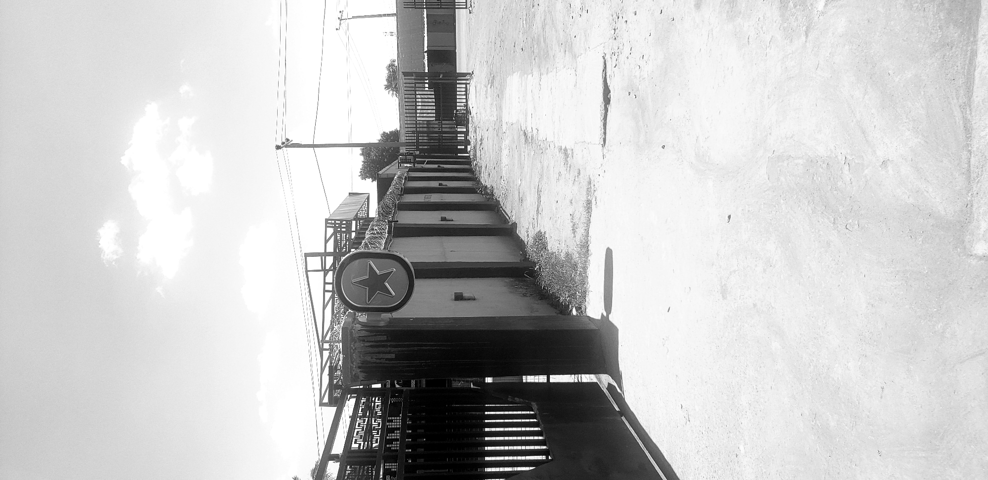 This black and white photograph captures an outdoor scene in Narayi, Nigeria, depicting a paved pathway alongside a wall and gates under bright daylight with scattered clouds. The foreground consists of a light-toned, worn concrete or asphalt surface with visible cracks and minor debris. A large, dark metal gate, possibly made of vertical bars with decorative patterns, is partially open on the left, casting a dark shadow onto the ground.
Running along the right side of the paved area is a painted concrete or cinder block wall, featuring darker vertical structural elements. Attached to one of these darker elements in the midground is a prominent circular sign displaying a five-pointed star within its center. The top of the wall is fitted with coiled barbed wire or razor wire, indicating security measures. Behind the gate on the left, intricate metalwork and structural elements of a building or canopy are visible. Further down the path, a second, dark metal gate, similar in style to the first, is also partially open on the right side of the frame.
The background shows a bright sky with white cumulus clouds. Multiple utility poles supporting power lines stretch across the upper midground. Sparse vegetation, primarily weeds, grows at the base of the wall. No individuals are present in the scene, and no discernible actions are taking place. The overall impression is of an entrance or pathway to an industrial or commercial property, or a secured residential compound.