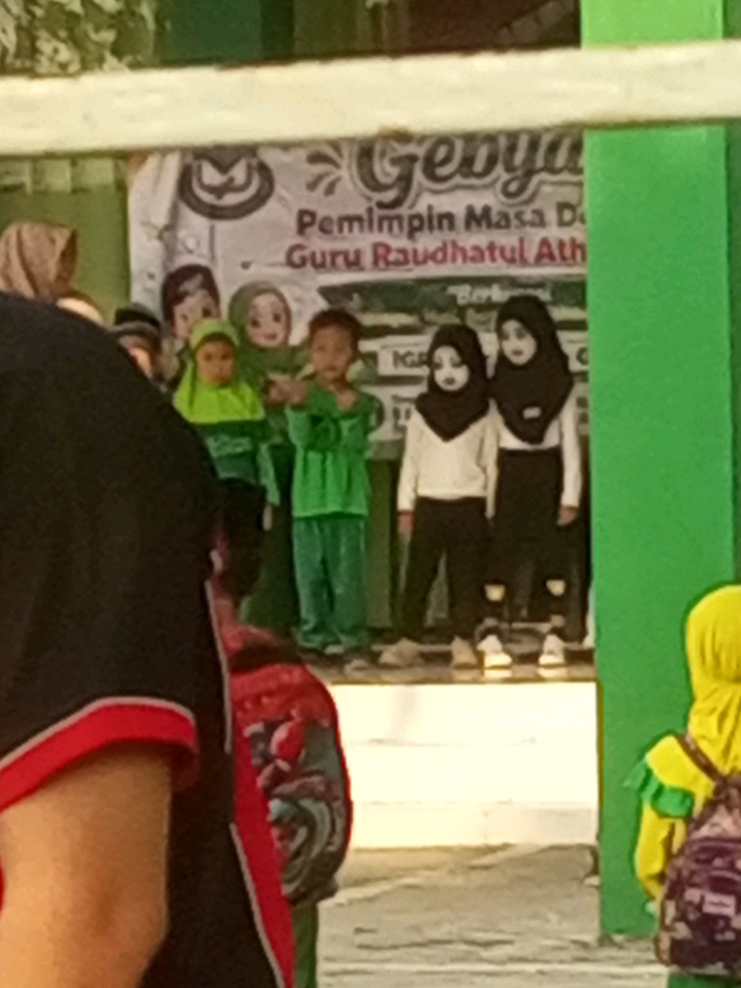 A group of children, likely students, are gathered outdoors in front of a banner. Some of the children are wearing green uniforms, including hijabs. Two girls in the foreground are wearing black hijabs and white tops with black pants. The banner behind them features text and cartoon-like illustrations of children's faces. The overall setting appears to be a school or community event in Gurah, Indonesia, suggested by the green building structures and the Indonesian text on the banner. The lighting indicates it is daytime.