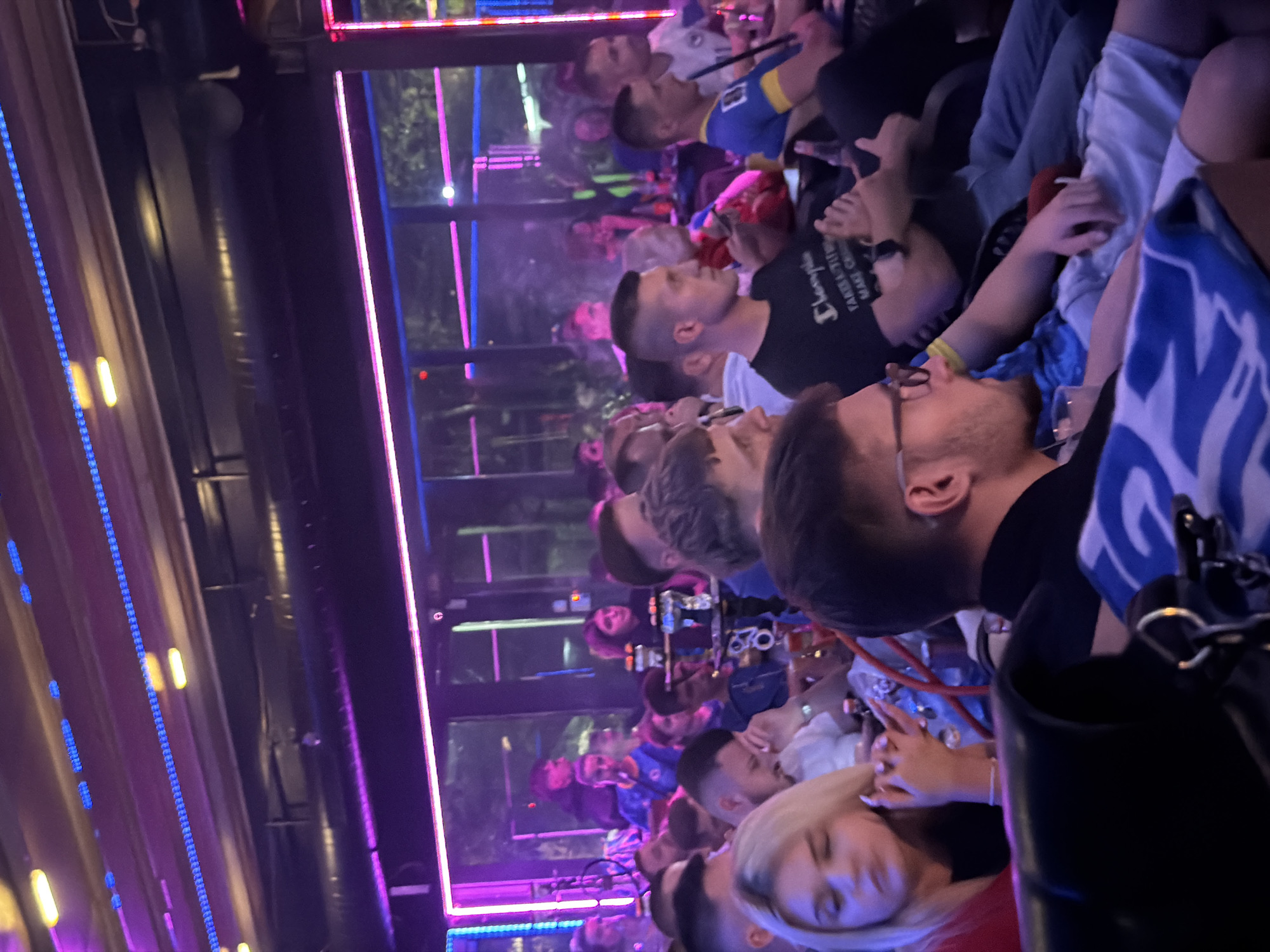The image shows a group of people gathered indoors, likely at a social venue such as a bar or club. The environment is dimly lit with prominent neon lighting, featuring blue and pink hues that illuminate the ceiling and some walls. Several individuals are visible, some in focus and others blurred, suggesting a candid shot or motion blur. Many appear to be seated, with some engaged in conversation or watching something. There are hookahs present, indicating a shisha lounge or a venue where smoking is permitted. The people are dressed in casual attire. The overall atmosphere suggests a relaxed social gathering. No specific landmarks or details within the image definitively place the scene in Travnik, Bosnia and Herzegovina.