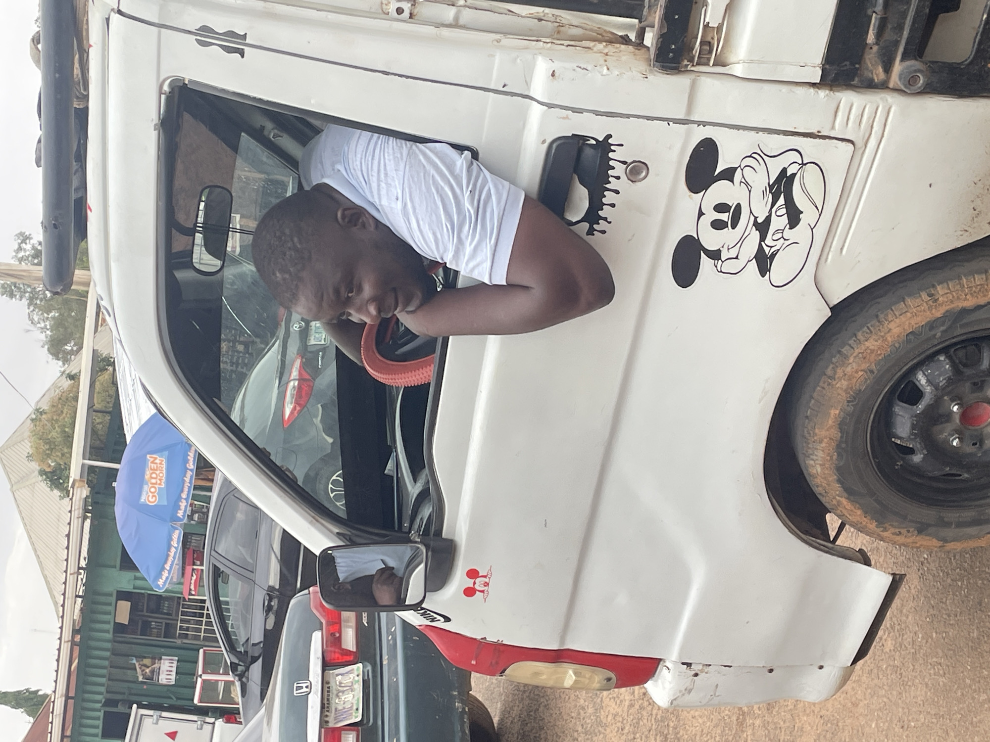 A man in a white t-shirt is leaning out of the window of a white truck parked on a city street in Jos, Nigeria. A Mickey Mouse decal is on the truck door. The environment includes other cars, a store with a blue "Golden Morn" umbrella, and greenery in the background.