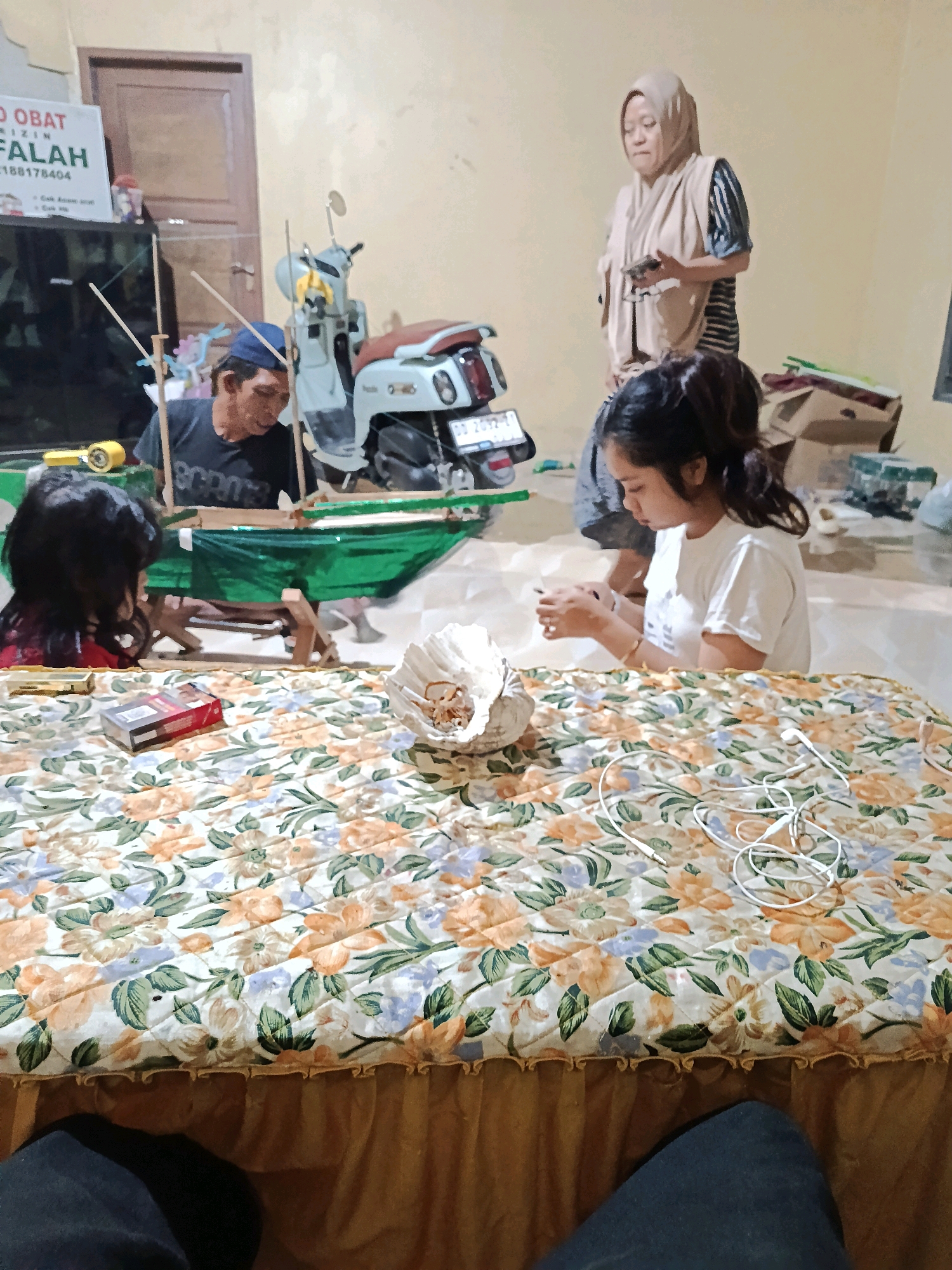 The image depicts an indoor scene in Bulukumba, Indonesia. In the foreground, a bed is covered with a floral patterned quilt, with a woman sitting on the right side of the bed, looking down at her hands and holding a mobile phone. To her left, another woman is also looking down. On the bed, there is a pack of cigarettes and a small bowl of what appears to be dried food. In the background, a man wearing a blue cap is working on a green object supported by wooden structures. Behind him, a motorcycle is parked. A woman in a headscarf stands in the background, looking at her phone. The environment appears to be a domestic or workshop setting.