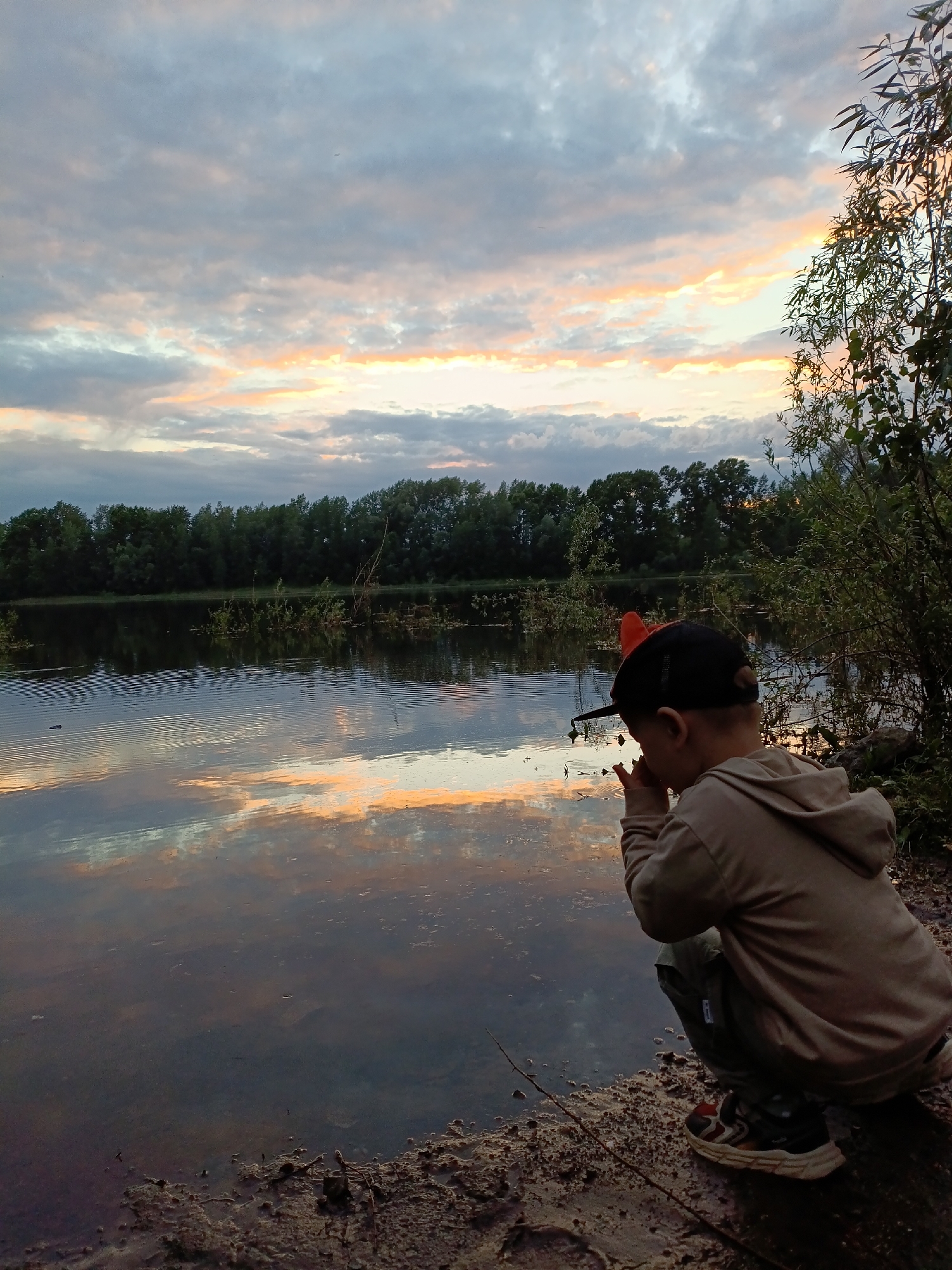 An evening scene unfolds at the Kama River embankment in Chaikovsky, Russia. A young boy, viewed from behind, is crouched down near the water's edge. He is wearing a tan hoodie, dark pants, a black baseball cap with an orange accent, and multi-colored sneakers. The boy appears to be focused on something near the water. The river reflects the dramatic colors of the sky, with a mix of grey, white, and soft orange clouds. The far bank of the river is lined with a dense row of green trees. The foreground consists of muddy, uneven ground with some sparse vegetation.