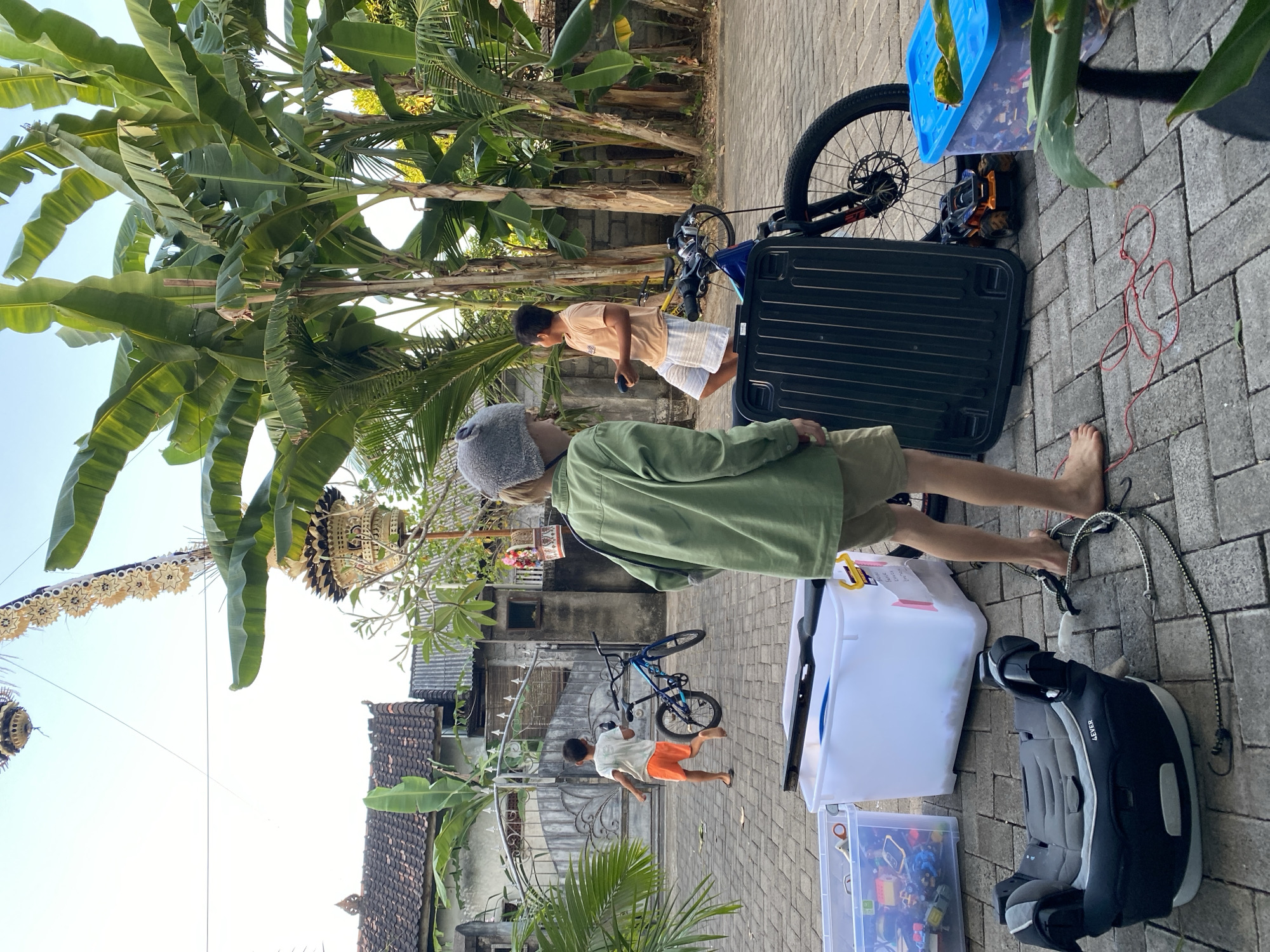 The image depicts a street scene in Kuta Selatan, Indonesia, likely in a residential area. Two young boys are present. One boy, wearing an olive green jacket and khaki shorts, is barefoot and facing away from the camera, with a grey beanie on his head. He appears to be looking towards a blue bicycle leaning against a gate. The other boy, wearing an orange t-shirt and white striped shorts, is also barefoot and is further down the street, running towards the gate with a small object in his hand. In the foreground, there are several large plastic storage containers, one filled with toys, and a car seat. A black ribbed object, possibly a panel or lid, and a mountain bike are also in the foreground. Banana trees are prominent in the background, with their large leaves dominating the upper right portion of the image. The houses in the background have tiled roofs. The ground is paved with interlocking bricks. A decorative Balinese banner hangs from a pole on the right side of the image.