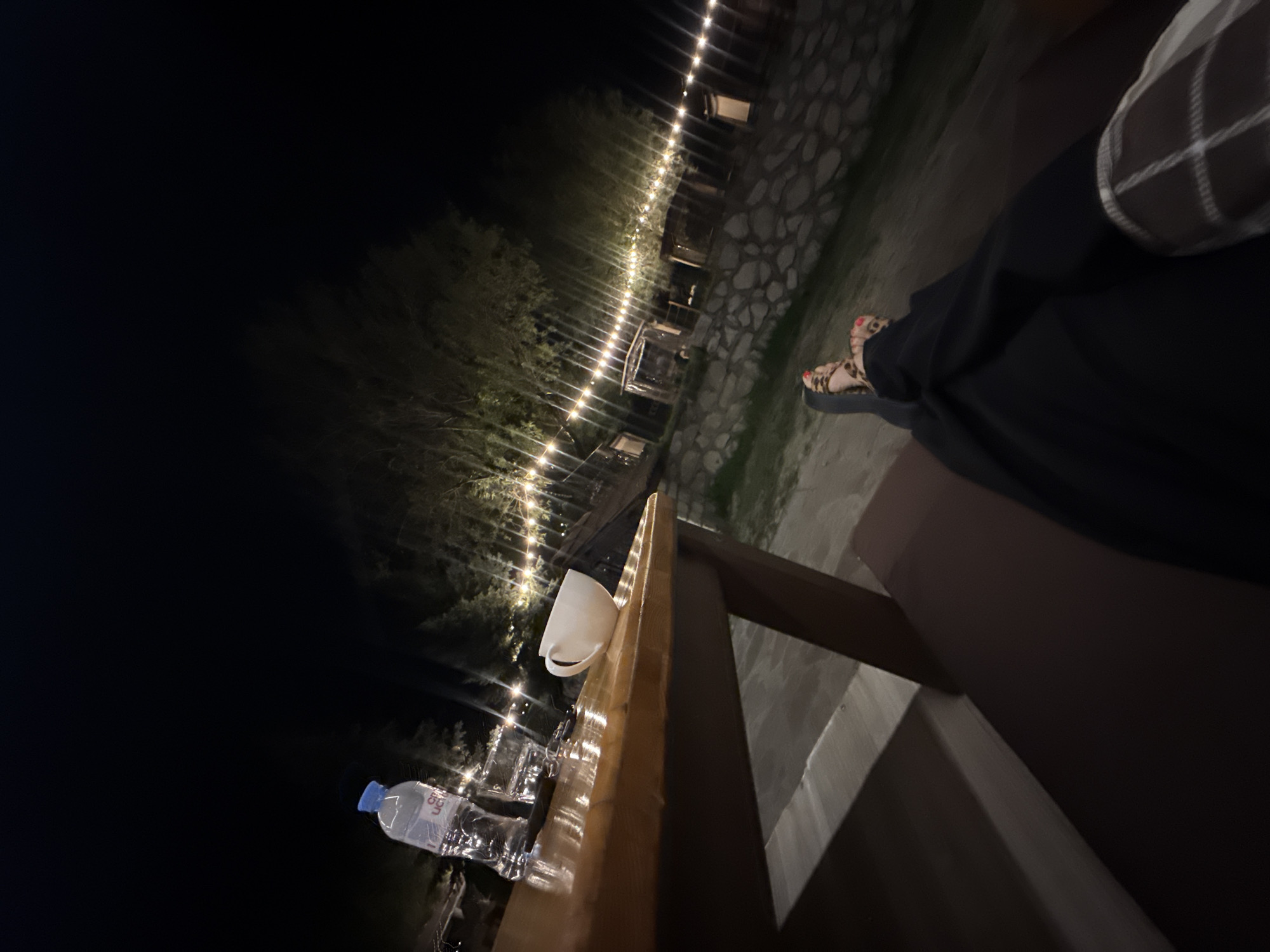 A person's legs and feet are visible in the foreground, resting on a dark surface. The person is wearing black pants and leopard-print sandals with red-painted toenails. The scene takes place outdoors at night, illuminated by a string of warm-toned lights that stretch across the background, creating a festive atmosphere. The lights are draped over trees and in front of several small structures, possibly sheds or enclosures, with a stone wall visible behind them. In the mid-ground, a wooden table holds a bottle of water and a white mug. The table's surface is polished and reflects the ambient light. The surrounding environment appears to be a rural or semi-rural area with trees and a dimly lit ground. The image suggests a relaxed outdoor setting, possibly a cafe or a gathering place, during nighttime hours. The specific location within Орлиновский округ, Ukraine is not discernible from the visual information alone.