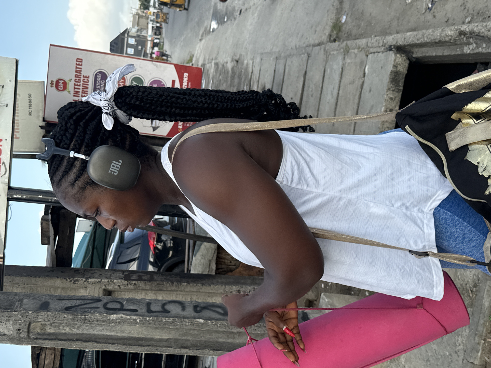 A medium shot depicts a woman wearing a white tank top, JBL headphones, and braided hair with a white bandana, standing on a concrete sidewalk. She carries a pink yoga mat and a bag with a gold design on her shoulder. In the background, there are parked cars, a street scene with distant figures, and commercial signs reading "GM INTEGRATED SERVICE," "RING STORE/," and "RC 1188850." The scene appears to be set in the city of Ado, Nigeria. The woman is looking down at something in her hands. The lighting is bright, suggesting an outdoor daytime setting.