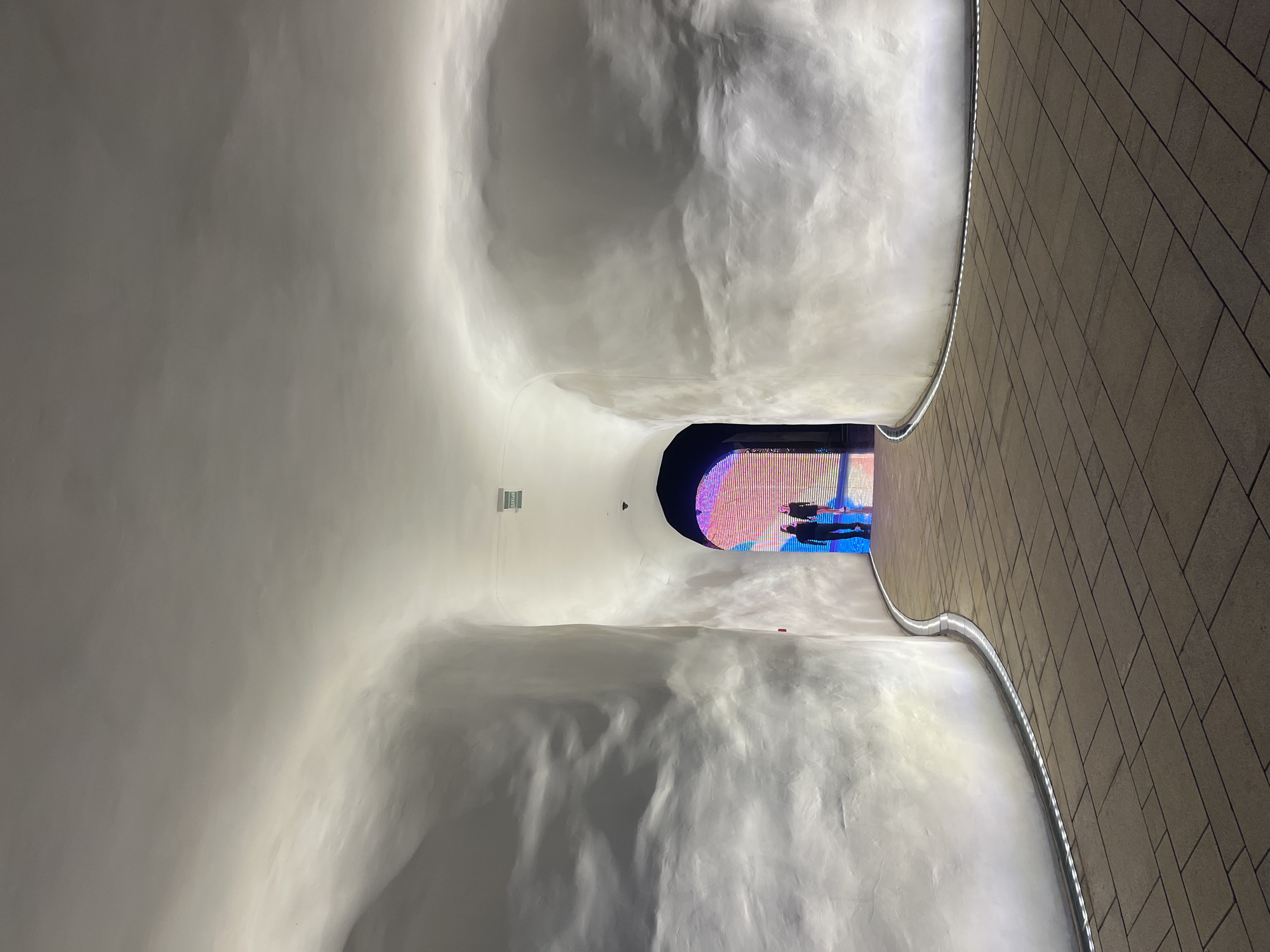This image captures an indoor scene with striking architectural features. Two individuals are visible in the distance, silhouetted against a brightly lit, colorful display or screen that fills the end of a tunnel-like passageway. The passageway itself is characterized by smooth, undulating white walls that create a soft, organic feel. On the right side of the image, a textured wall made of rectangular tiles runs parallel to the curved passageway, accented by a subtle strip of lighting along its base.
The environment appears modern and potentially artistic or exhibition-like, given the dynamic lighting and the large illuminated display. The lighting suggests it might be evening or an indoor space where artificial light is dominant. The overall atmosphere is one of visual interest, with the contrast between the organic curves of the white walls and the geometric pattern of the tiled wall. The presence of the people at the end of the tunnel draws the viewer's eye towards that focal point, hinting at an area of interest or an exit. There is a small red object, possibly a marker or piece of art, on the white wall in the mid-ground. A green sign with white text is also visible on the white wall in the upper left portion of the image, though the text is not legible. The location context is Krasnodar, Russia.