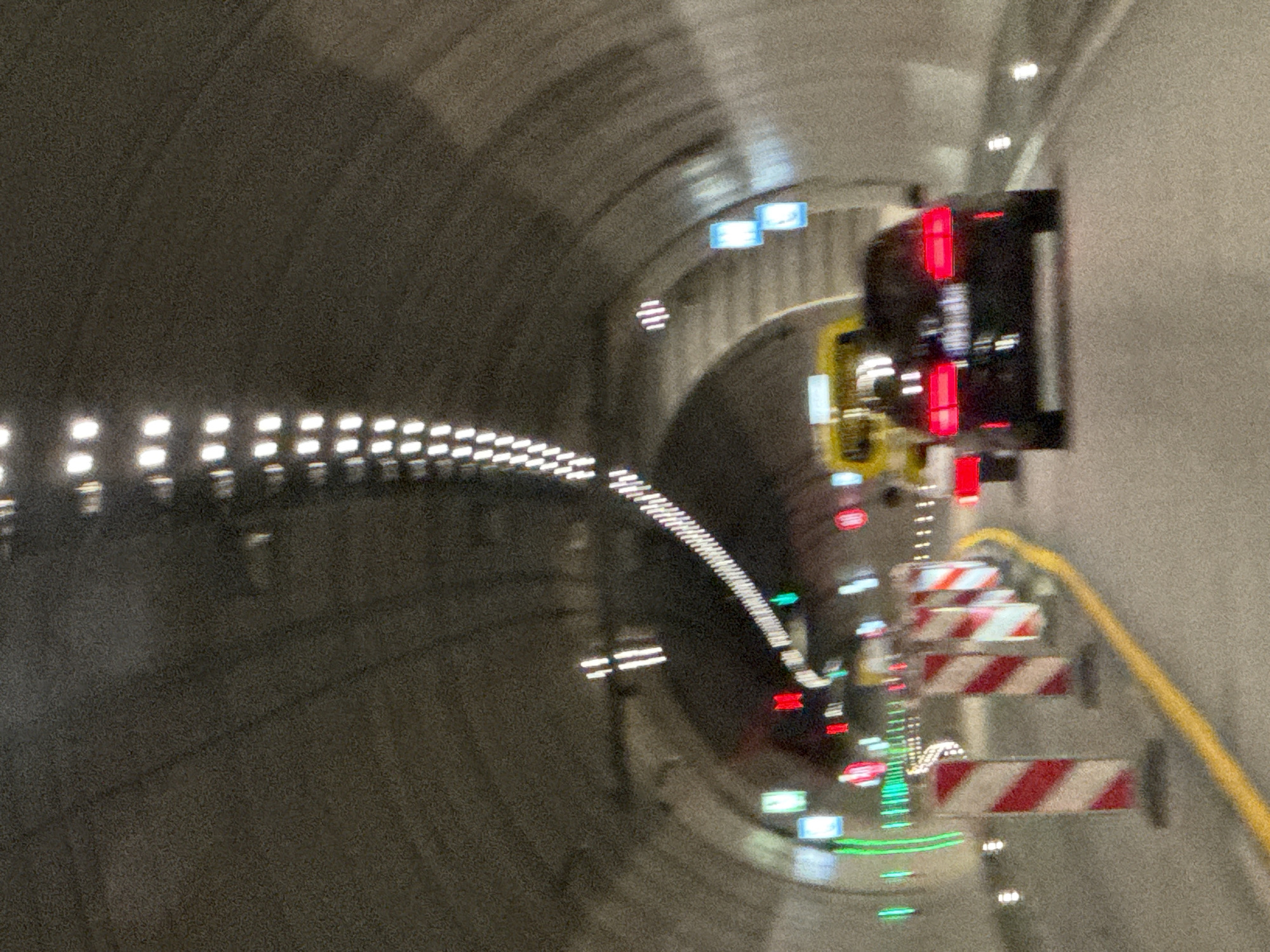 The image depicts a view from within a tunnel, likely part of the A4 motorway in Kodersdorf, Germany, given the context provided. The tunnel is illuminated by rows of white lights along the ceiling, creating a somewhat blurred, streaky effect due to motion blur from the camera. The road surface is asphalt, with a distinct yellow line marking the right lane.
In the foreground, several striped traffic barriers, red and white, are positioned along the side of the road, suggesting some form of construction or lane closure. Further ahead, a yellow bus or similar large vehicle is visible. Behind the bus, a dark-colored car with its red taillights illuminated is traveling in the same direction. Several illuminated signs are mounted on the tunnel walls, some appearing green and others blue, their exact purpose unclear due to the blur. The tunnel itself has a concrete, arched structure. The overall scene conveys a sense of travel through an enclosed, man-made passage, likely during nighttime or low-light conditions.