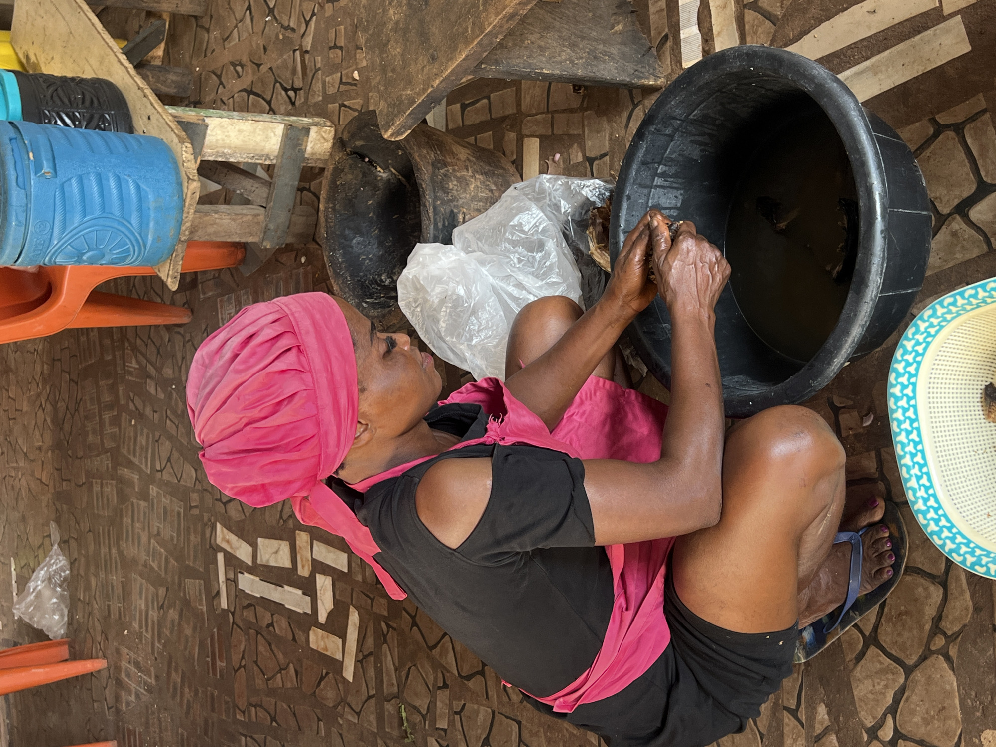 In Nkpor, Nigeria, a woman wearing a pink headwrap and a black dress with a pink apron is kneeling on a patterned tiled floor. She is in the process of preparing food, with her hands submerged in a large black basin filled with water and pieces of fish. To her right, a white plastic bag is visible, and behind her, a large dark pot sits on the floor. In the background, an orange plastic chair and a wooden bench with blue and yellow containers are present. The scene appears to be an outdoor or semi-open food preparation area.