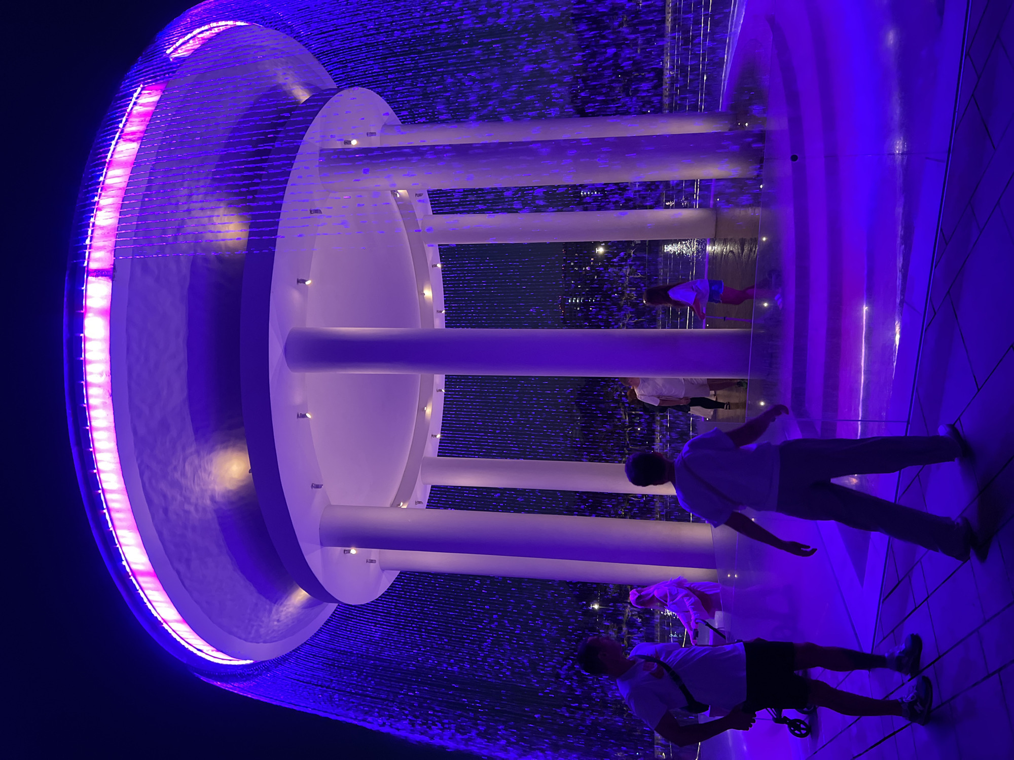 This media file captures an illuminated water feature with people present. The scene is dominated by vibrant purple and pink lighting that reflects off the water and the surrounding architecture. The water appears to be falling in thin streams, creating a curtain-like effect.
The primary subjects are several individuals. In the foreground, two men are visible, one wearing a white t-shirt and light-colored pants, the other in a white t-shirt and dark shorts. They seem to be observing or interacting with the water feature. Further in the background, a person in a pink top and another in a white shirt are also present.
The setting appears to be an outdoor public space, likely an urban environment, given the tiled walkway and the visible architecture. The lighting suggests it is nighttime or dusk. The overall mood is one of visual spectacle and perhaps a relaxed atmosphere, as people are casually present.
Notable details include the circular structure of the water feature, with white cylindrical columns supporting layered platforms. The dynamic flow of the water, illuminated by LEDs, is a key visual element. There is no visible text in the image. The location context of Krasnodar, Russia, is provided but not visually indicated within the image itself.