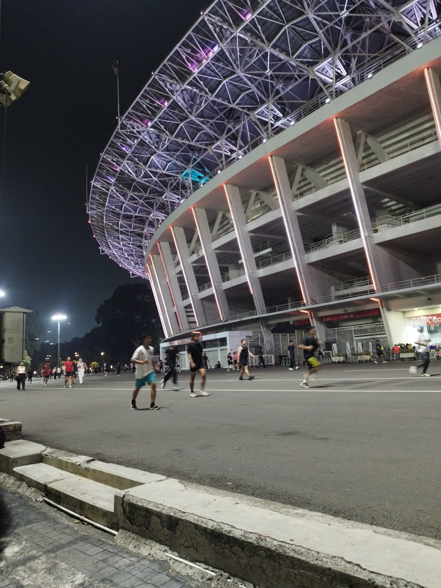 At night, the exterior of a large stadium, illuminated by a mix of purple and orange lights, curves into the frame from the upper right. The stadium features a complex metal superstructure and multiple levels of concrete seating. In the foreground, a paved road runs horizontally across the image, with a concrete curb and steps to its left. A group of people, mostly in athletic wear, are walking and running on the road. Some individuals are in focus, appearing to be mid-stride or in conversation, while others are blurred due to motion. The background is dark, with the silhouette of trees and distant streetlights visible. The scene appears to be in an urban environment, likely an area surrounding a sports complex in Central Jakarta, Indonesia, possibly Gelora Bung Karno Stadium, given the architectural style and the presence of people engaging in evening exercise.