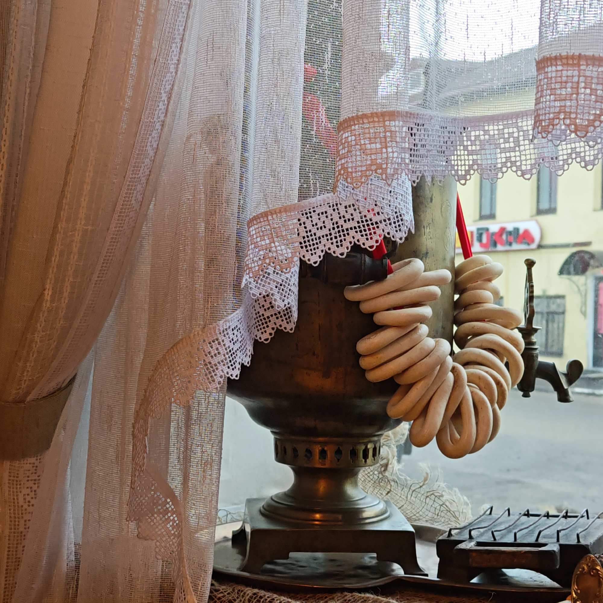 The image shows a samovar decorated with white lace and strings of bagels, sitting by a window with white lace curtains. Outside the window is a view of a building, possibly in Rostov, Russia.