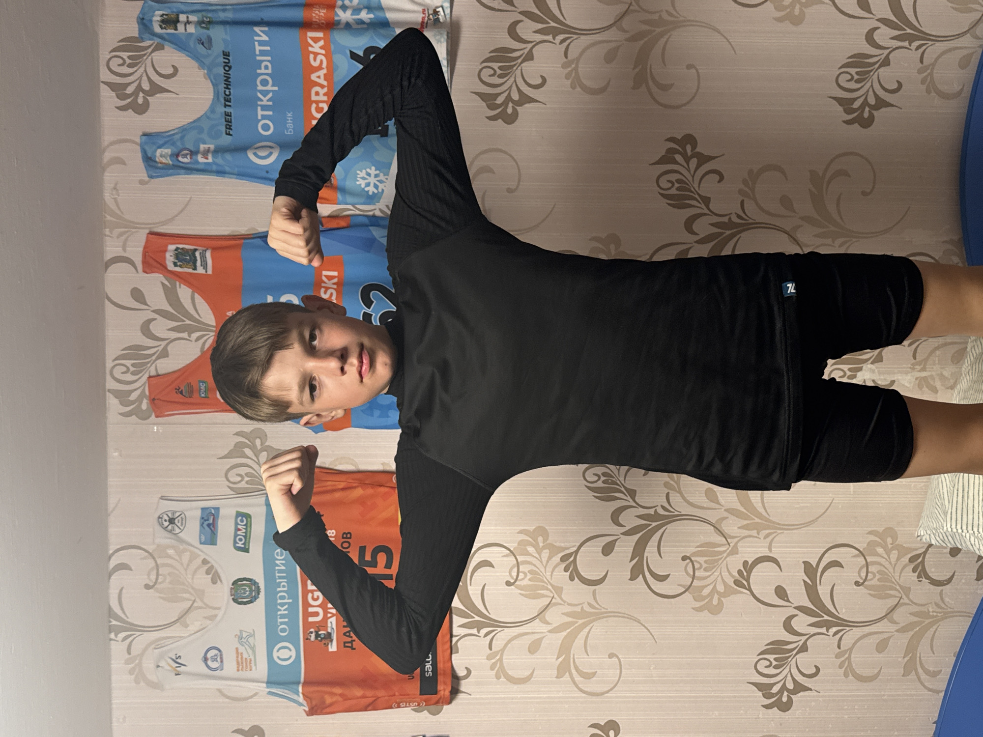 The image shows a young boy standing against a wall with patterned wallpaper. He is wearing a black long-sleeved shirt and black shorts, and he is flexing his arms. Behind him on the wall are four sports jerseys. The jerseys contain the word "GRASKI" and Cyrillic text that translates to "Opening". One jersey also contains "SKI". Other texts present on the jerseys are "FREE TECHNIQUE", "ЮМС", "ДАН", "UG" and "ЮМС". One jersey features the number 15 and the name of the city Chaikovsky, as well as the name "OV". One jersey bears the acronym "F/S". The jersey also includes the Russian words "Открытие", and "банк". The image was likely taken in Chaikovsky, Russia.