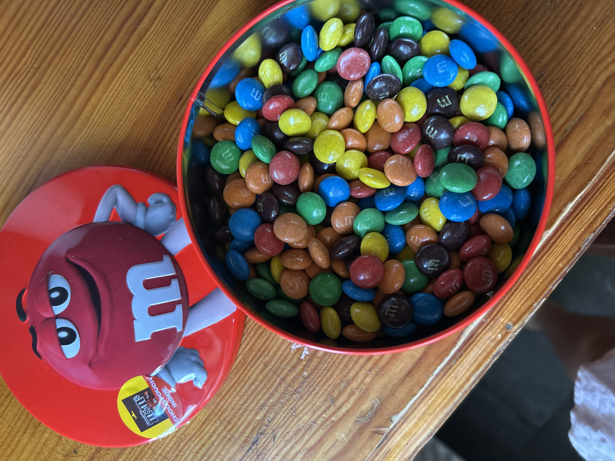 The image shows a red tin of M&M candies on a wooden table. The tin lid, featuring a cartoon M&M character, is partially open, revealing a colorful assortment of M&M candies inside. The candies are of various colors including red, blue, green, yellow, brown, and orange. The table surface has a visible wood grain pattern. The image does not contain any information about the city of Basarabeasca, Moldova, or any people.
