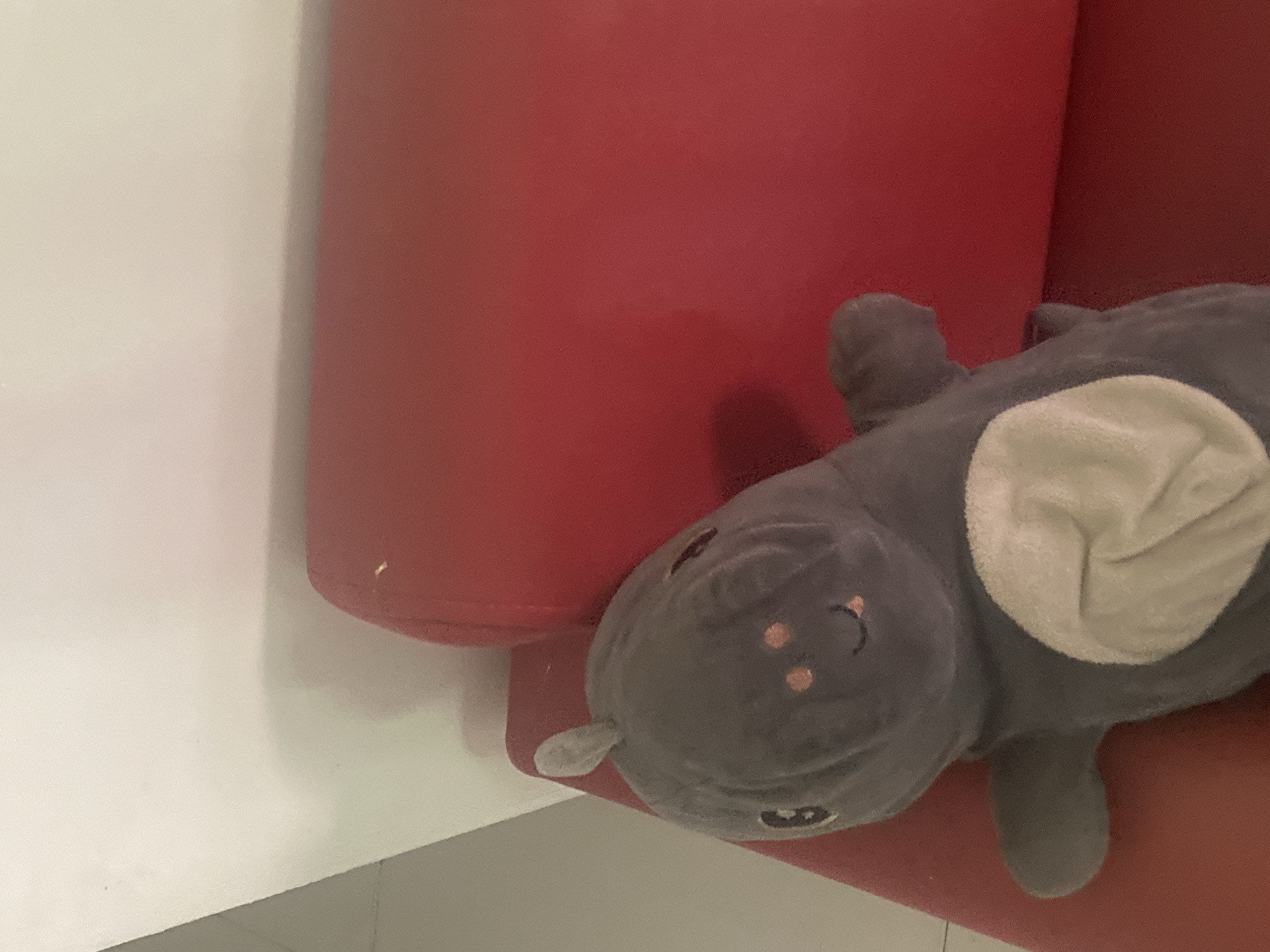 A stuffed animal, which appears to be a gray hippopotamus, rests on a red sofa. The hippopotamus is positioned on its side, with its head turned towards the camera. It has a white belly patch and embroidered facial features, including eyes, a mouth with a tongue, and two pink cheeks. A small grey ear is visible on the top of its head. The red sofa is the primary object in the foreground, with a portion of a white wall visible in the upper left corner. The scene is indoors, and there is no indication of it being in Proyonangg, Indonesia, or any other specific city.