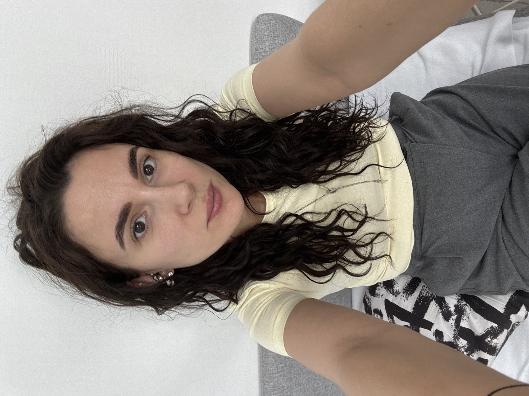A young woman with long, dark, wavy hair is depicted taking a selfie indoors, appearing to be lying down or leaning back. She has dark eyes and eyebrows, with a neutral expression and slightly parted, light pink lips, looking directly into the camera. She wears a light yellow, long-sleeved top and dark grey trousers. On her visible left ear, she has several small, metallic earrings, and a thin silver chain is visible around her neck.
The background consists of a plain white surface, possibly a wall or bed sheet, on the left side. Behind her head and shoulders, a light grey upholstered surface, likely part of a sofa or bed, is visible. To the right, a black and white patterned pillow or blanket, featuring bold, abstract brushstroke designs, adds a visual accent. The overall lighting is bright and even, indicating a daytime setting. There is no visible text in the image. The location context for this media file is Chaikovsky, Russia.