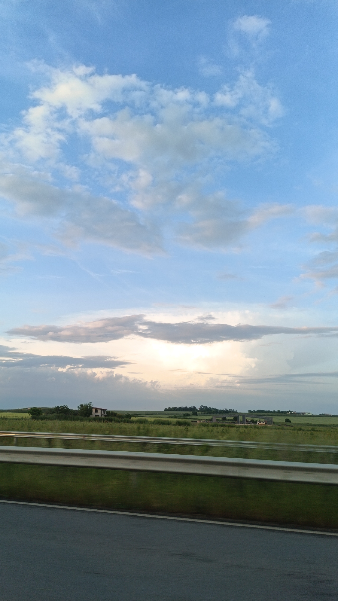 The image depicts a view from a moving vehicle, likely on a highway or major road, with a clear blue sky and scattered clouds above. In the middle ground, there's a field of green vegetation with a small house or building visible among the trees and bushes. A metal guardrail runs along the side of the road. It's not possible to determine the location in Burgas, Bulgaria, from this single image without further contextual information.