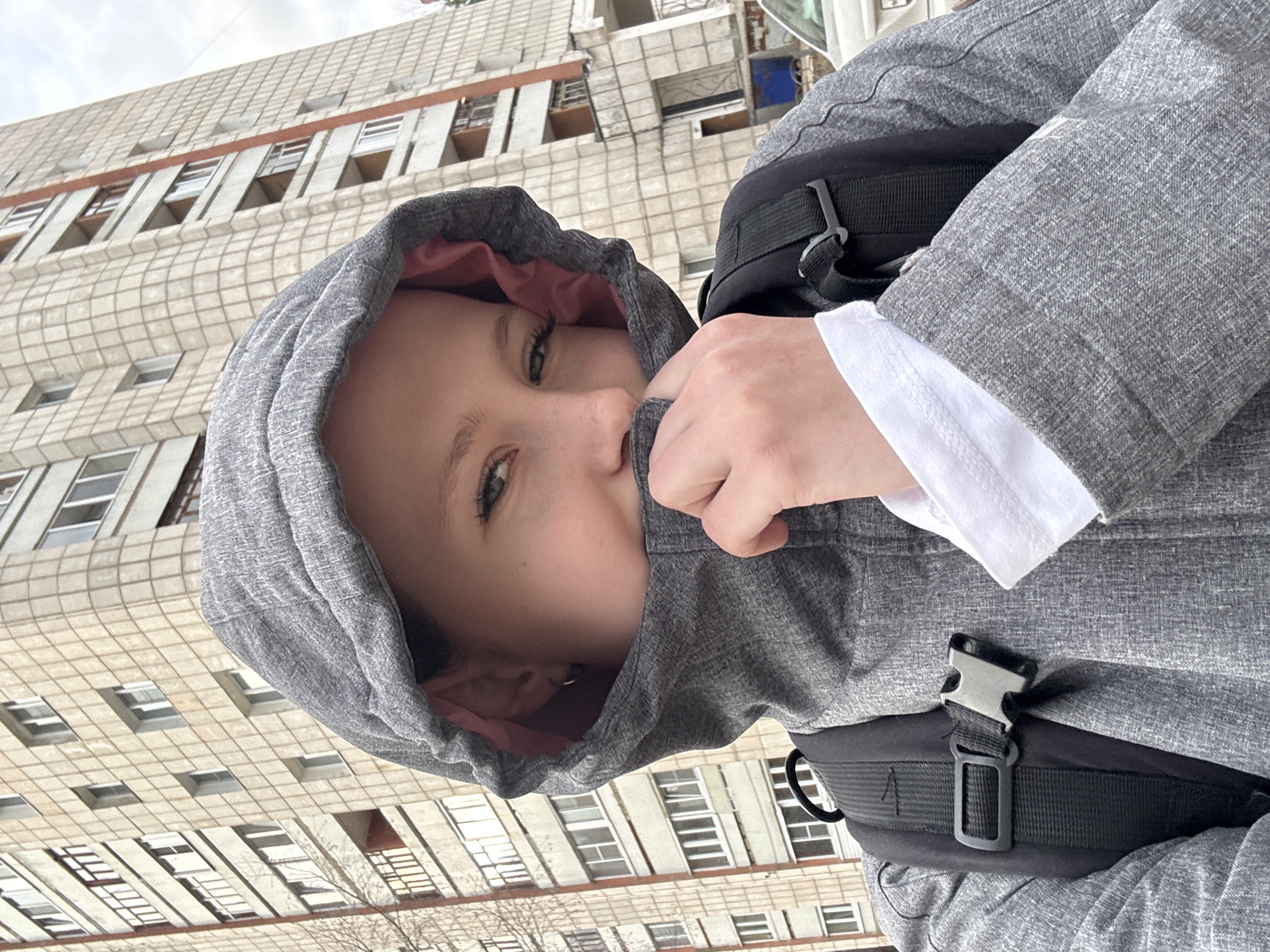 A close-up portrait features a young person, likely female, wearing a grey hooded jacket with a visible pink lining. Their left hand pulls the front of the jacket up to cover their mouth and nose, leaving their eyes looking directly at the camera. A white cuff of a shirt or undershirt peeks out from under the jacket's right sleeve. A black backpack or bag strap with a silver buckle is visible over their right shoulder.
The background is dominated by a large, multi-story building with a distinctive grid-patterned facade, likely made of light-colored concrete or tiles. Numerous rectangular windows, some recessed, are visible across its surface, along with what appear to be small, exposed balcony structures on some floors. The architectural style suggests a mid-20th-century urban structure, typical of locations such as Chaikovsky, Russia. The sky overhead is overcast and uniformly grey, indicating a cloudy day. The person's attire and gesture of pulling up the hood suggest cool or cold weather. No visible text is present in the image.