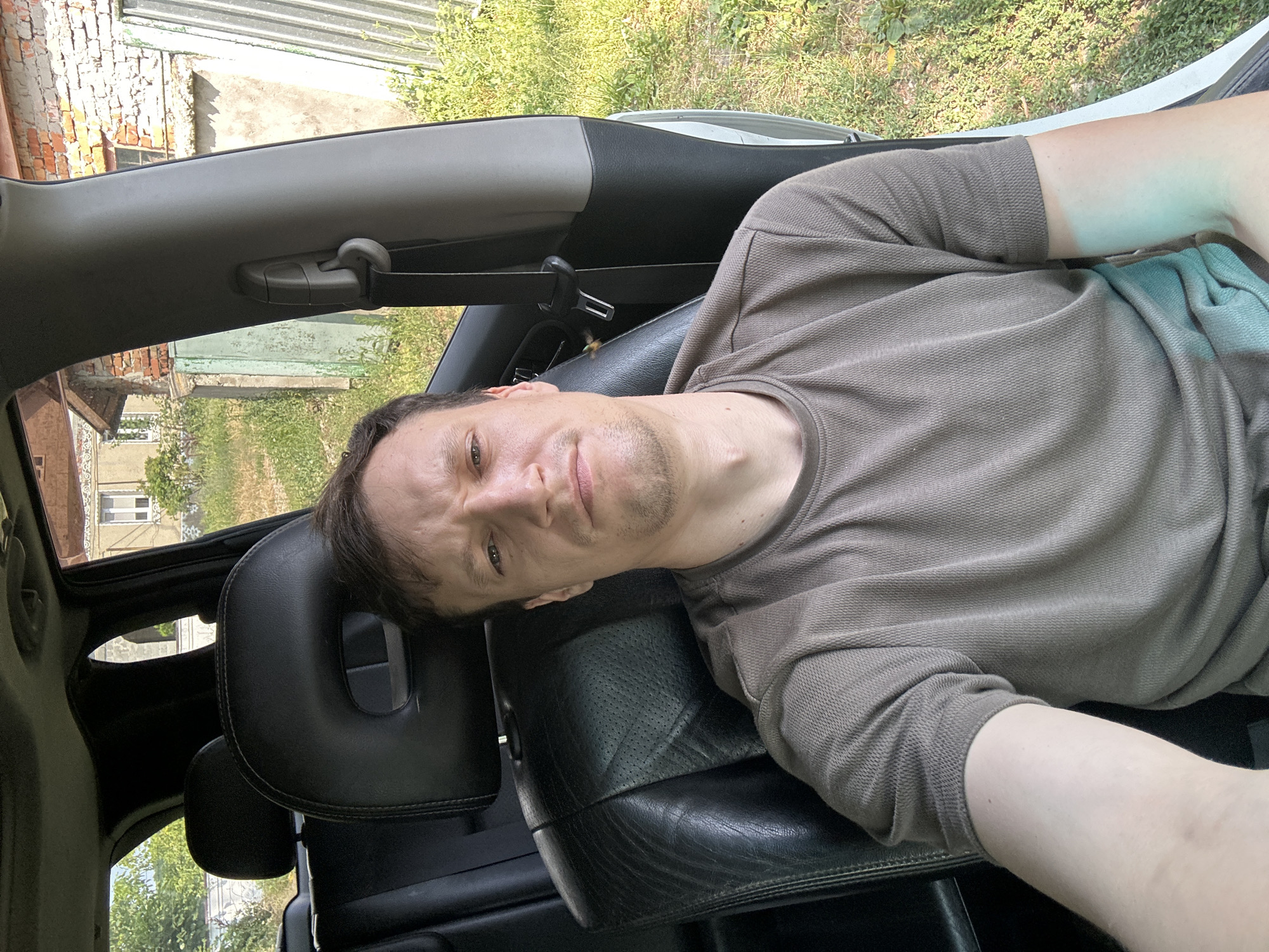 A medium shot selfie depicts a fair-skinned male with short brown hair and a light stubble, wearing a plain brown t-shirt, sitting in the driver's seat of a car. The man's face is directed towards the camera with a neutral expression, his brow slightly furrowed. The car's interior is visible, with black leather seats and a dark headrest. A grey seatbelt mechanism is attached to the car's pillar to the right of the man. Outside the car, a rural setting is observed, characterized by green grass, weeds, a portion of a brick building with a corrugated metal roof, and a partial view of a tall, light-colored building with a window. The background suggests an outdoor environment, possibly in a village or a less urbanized area. The timestamp indicates the image was taken in Makiv, Ukraine.