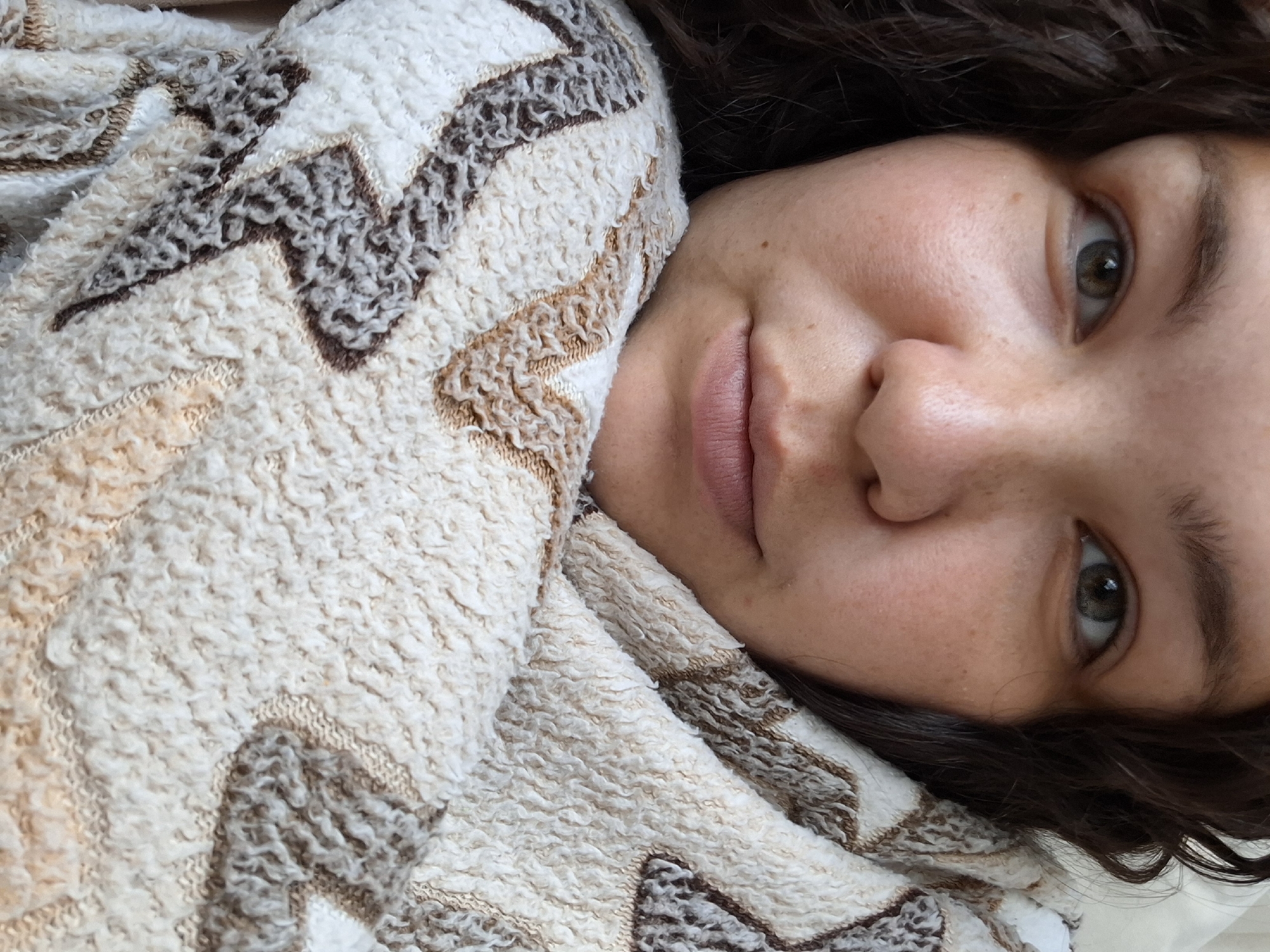 A close-up, rotated image features a person with brown hair, light skin, and green eyes, partially covered by a patterned blanket or towel. The blanket has a textured, looped surface with a geometric zig-zag design in beige, brown, and white. The person's face is in focus, showing details like freckles and lip texture. The image composition is intimate and portrait-like, focusing on the individual's facial features and the cozy texture of the blanket. The location in the city of Chaikovsky, Russia, cannot be determined from the image provided.