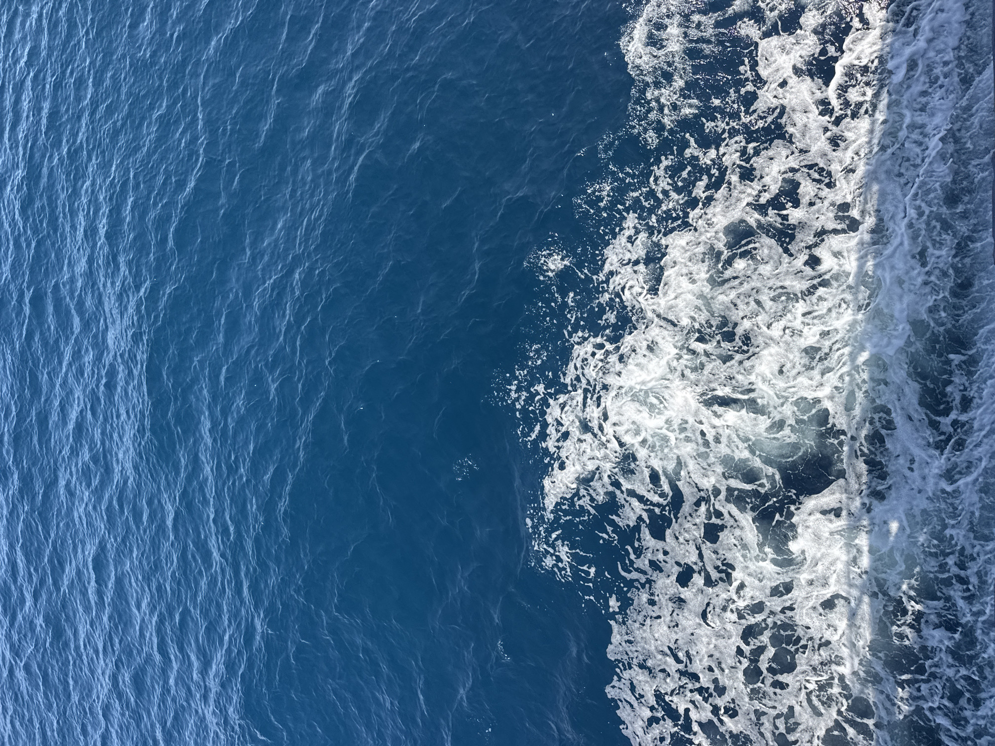 The image shows a close-up, high-angle view of the surface of deep blue water. The water is rippled, with a distinct line of white foam and turbulent waves in the foreground, suggesting movement, likely a boat wake. The background consists of calm, unbroken water. There are no discernible objects, people, or structures in the image, and the location is unknown.