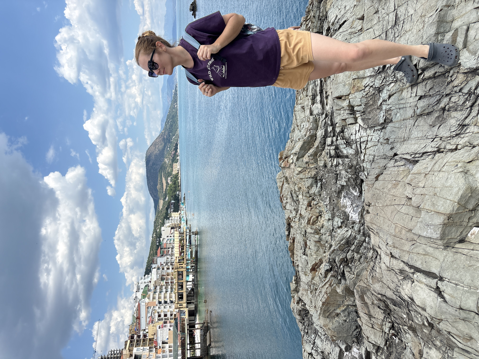 A blonde woman with her hair in a bun stands on a rocky outcrop overlooking a coastal town. She is wearing a purple t-shirt with a graphic, yellow shorts, and dark grey Crocs. She has a blue backpack on. The town, situated in Kiparisnoe, Ukraine, features a waterfront lined with multi-story buildings. The sea is a deep blue, and the sky is filled with white, puffy clouds. Mountains are visible in the distance.