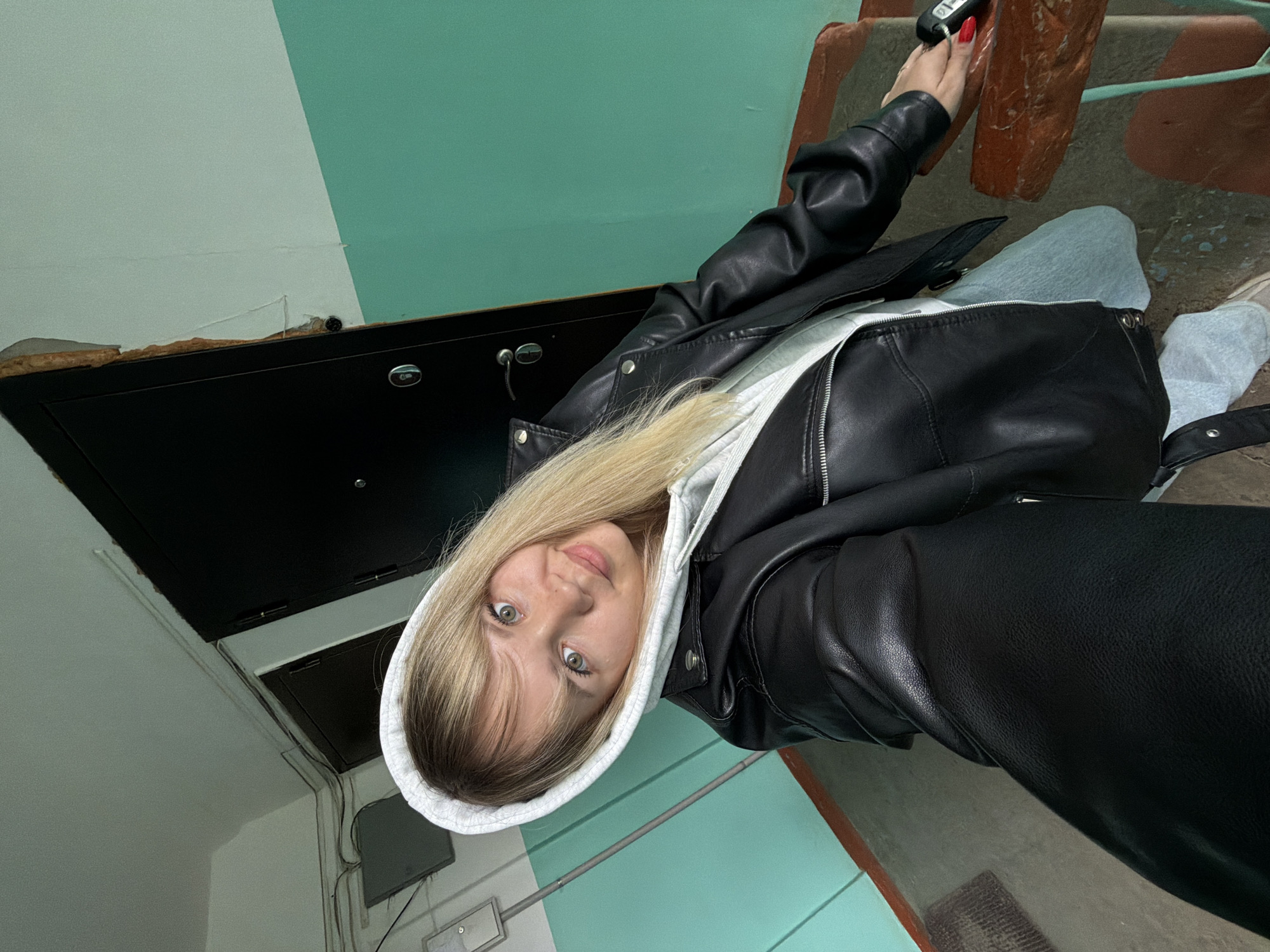 A young woman with blonde hair and green eyes is in a stairwell. She is wearing a white hooded sweatshirt and a black leather jacket. She is holding keys in her right hand. The walls are painted white and aqua. There is a black metal cabinet on the wall. The location may be an apartment building in Chaikovsky, Russia.