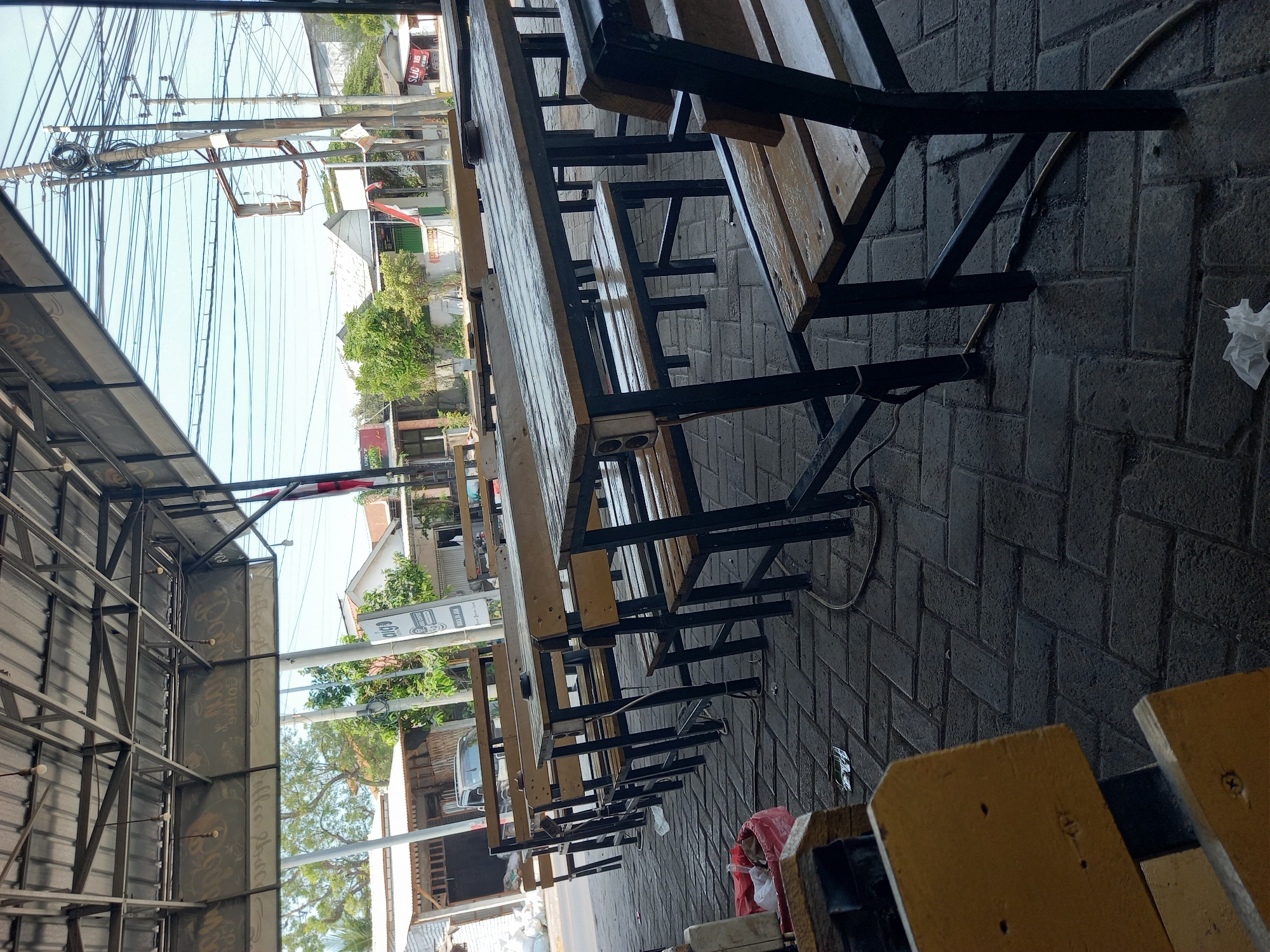 The image depicts an outdoor seating area at a food stall or restaurant on a street in Tunggul, Indonesia. The area features multiple long, wooden tables with matching benches, all constructed with black metal frames. The tables and benches appear to be simple and functional, designed for casual dining. The ground is paved with grey interlocking bricks.
In the background, a street is visible with parked cars and a few buildings that suggest a commercial or residential area. Overhead, a tangle of power lines crisscrosses the sky, a common sight in many Indonesian cities. The overall atmosphere is one of everyday life, with the seating area ready to serve customers. There are no people visible in the immediate seating area, suggesting it is either early in the day or a lull in activity. The lighting suggests it is daytime, with the sun casting shadows.