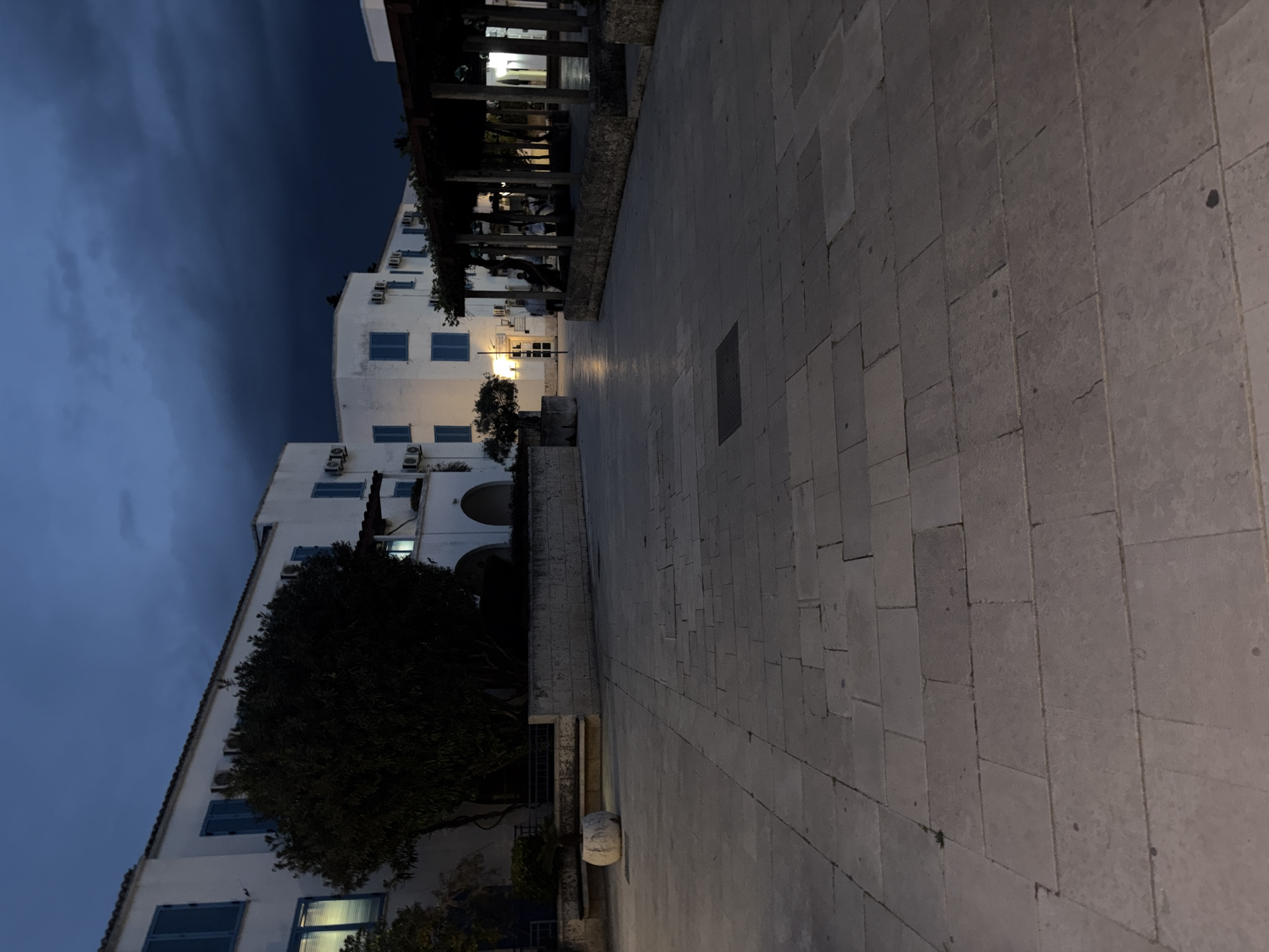The image shows a street scene in Budva, Montenegro, at night. The sky is dark blue, and the buildings are white with blue shutters. A stone-paved street leads away from the viewer, flanked on one side by a building and a tree and on the other side by a covered walkway supported by columns. There are no people visible in the image. A single light source illuminates a doorway on the building, while other lights shine from under the walkway. The air is still and the scene is dark.
