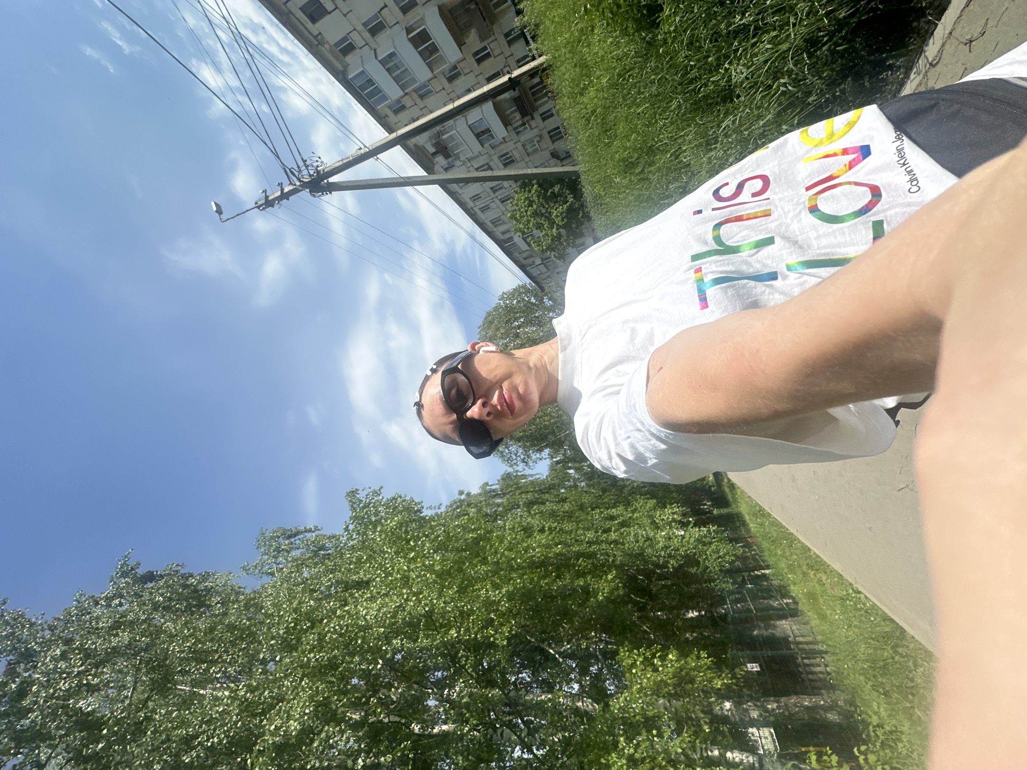 A woman wearing sunglasses and a white t-shirt is taking a selfie outdoors on a sunny day. The shirt has the words "THIS LOVE" printed on it in rainbow colors, and "Calvin Klein Jeans" printed on the shoulder. The background consists of trees, grass, a building, a utility pole, and a blue sky. The photo may have been taken in Votkinsk, Russia.