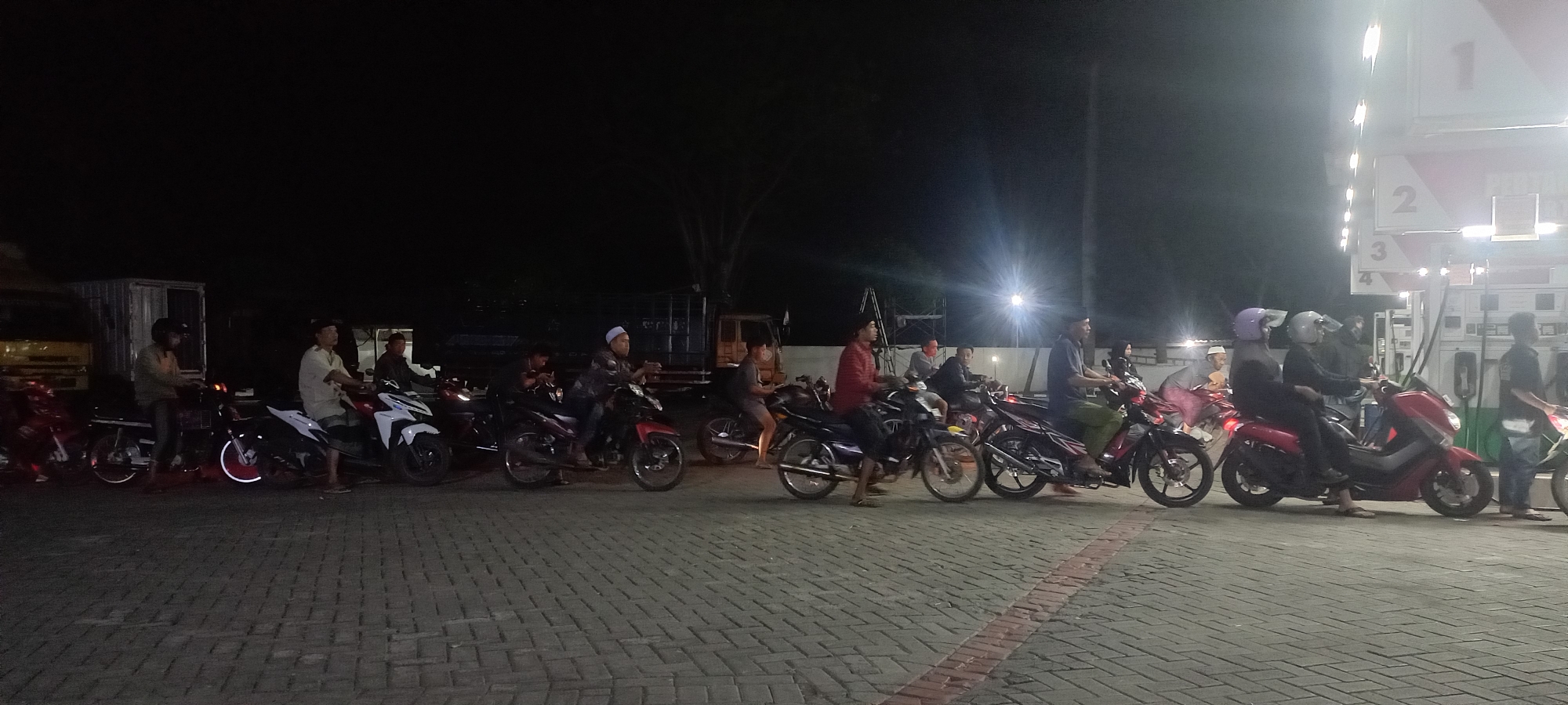 At night, a group of people on motorcycles are gathered at a gas station in Curahdringu, Indonesia. The scene is illuminated by artificial lights from the gas station. Several motorcycles are parked in a line on the paved ground, with people sitting on them or standing nearby. The gas station has visible pumps and signage. Some individuals are wearing helmets. The background is dark, suggesting it is nighttime.