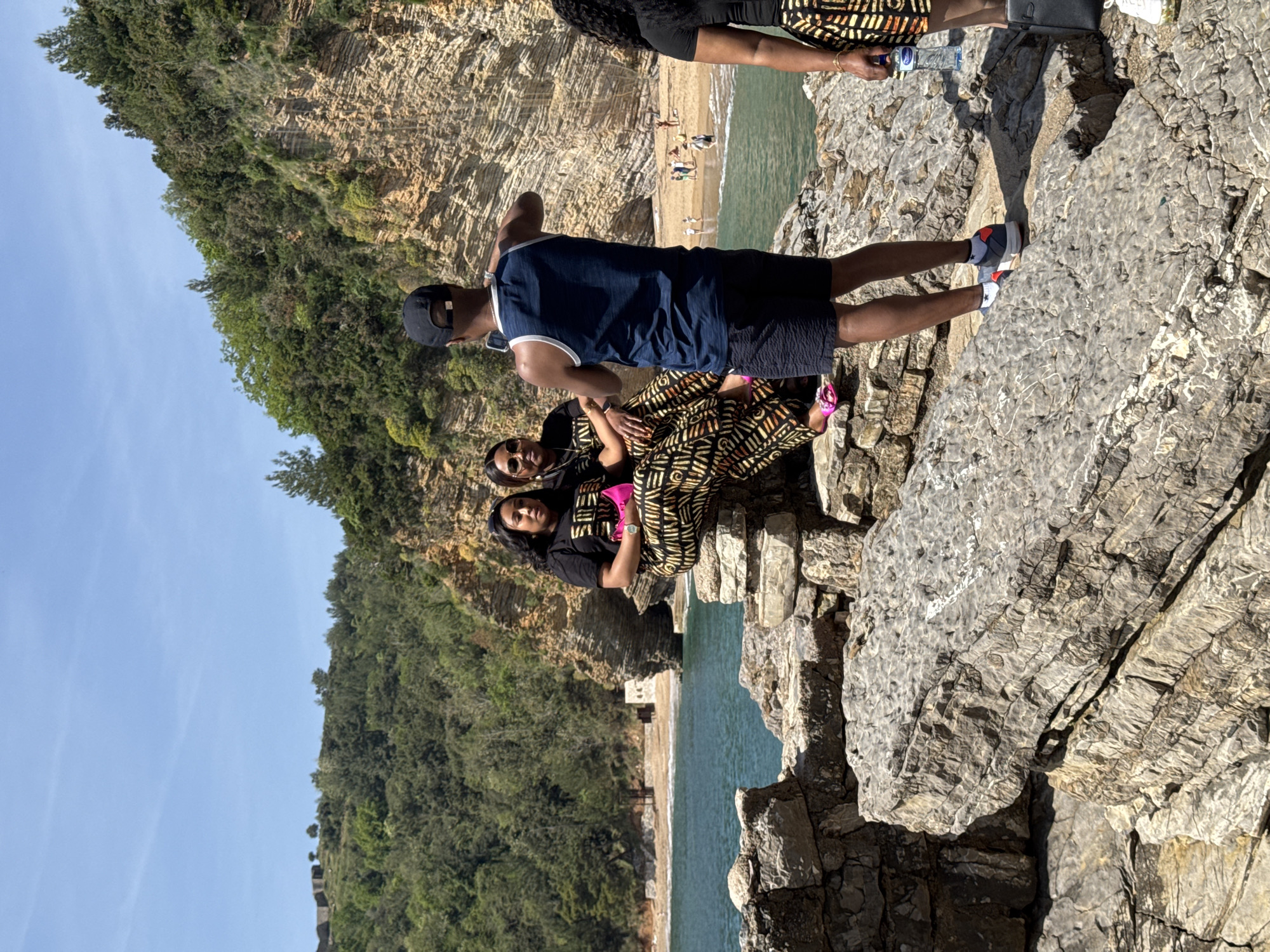 On a sunny day in Budva, Montenegro, a group of people pose for a photo on a rocky outcrop overlooking a beach. The group consists of three women and one man. The women are wearing black shirts and patterned skirts. The man is wearing a blue tank top and black shorts. The beach is populated with other people. The surrounding landscape includes rocky cliffs covered with greenery and a clear blue sky. A portion of the Adriatic Sea is visible. The location is possibly near Mogren Beach, given the coastal rock formations and beach proximity.