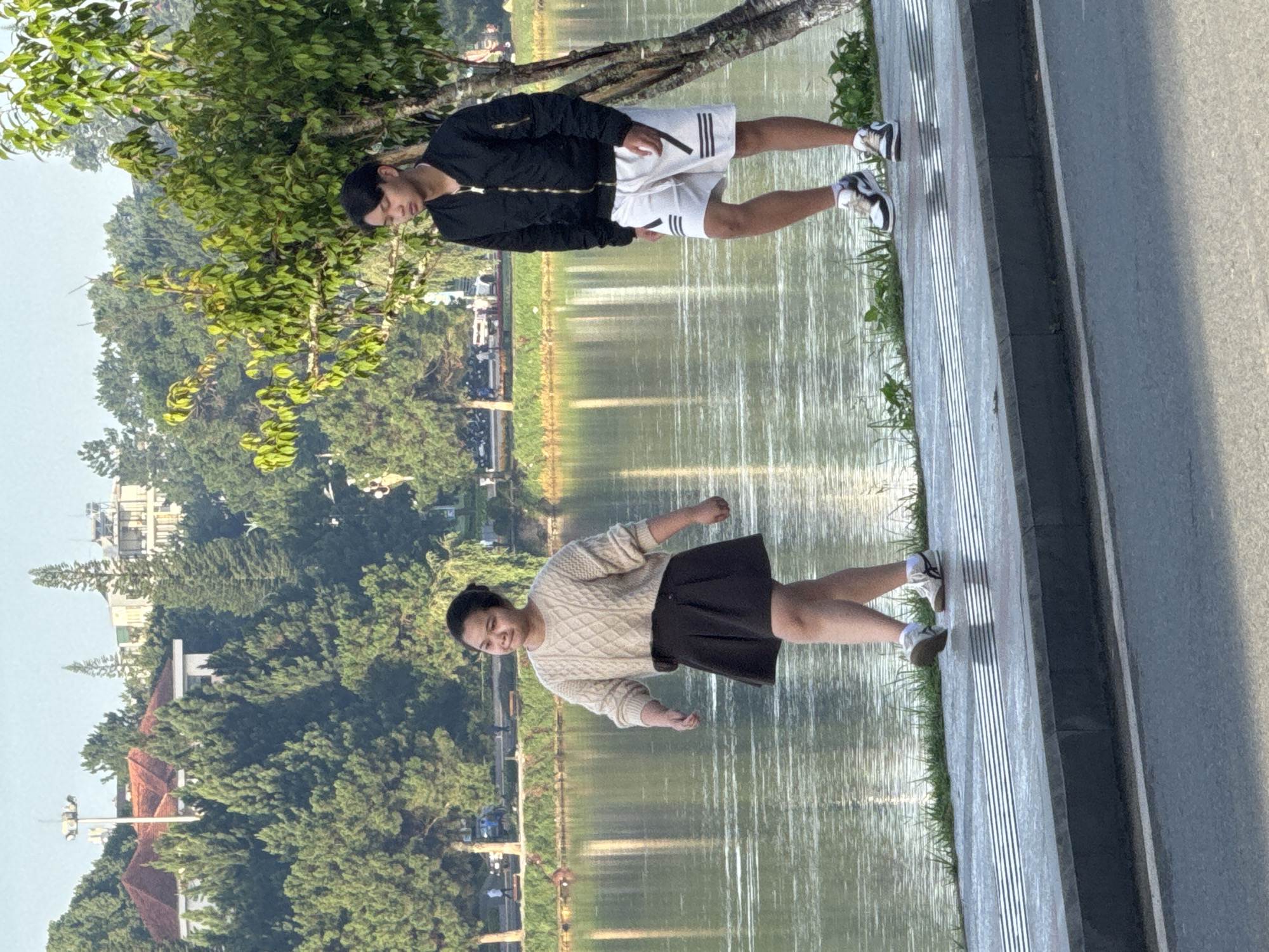 Two young people, a female and a male, are walking along a paved path next to a calm body of water. The female, on the left, wears a cream-colored cable-knit sweater and a dark, pleated skirt. The male, on the right, is dressed in a black bomber jacket and white athletic shorts with black stripes. They are both wearing athletic shoes and appear to be walking in the same direction. In the background, lush green trees cover rolling hills, and buildings are visible on the higher ground. The water reflects the trees and sky, creating a serene backdrop. This scene likely depicts Xuan Huong Lake in Da Lat, Vietnam, a popular location known for its scenic beauty and recreational pathways.