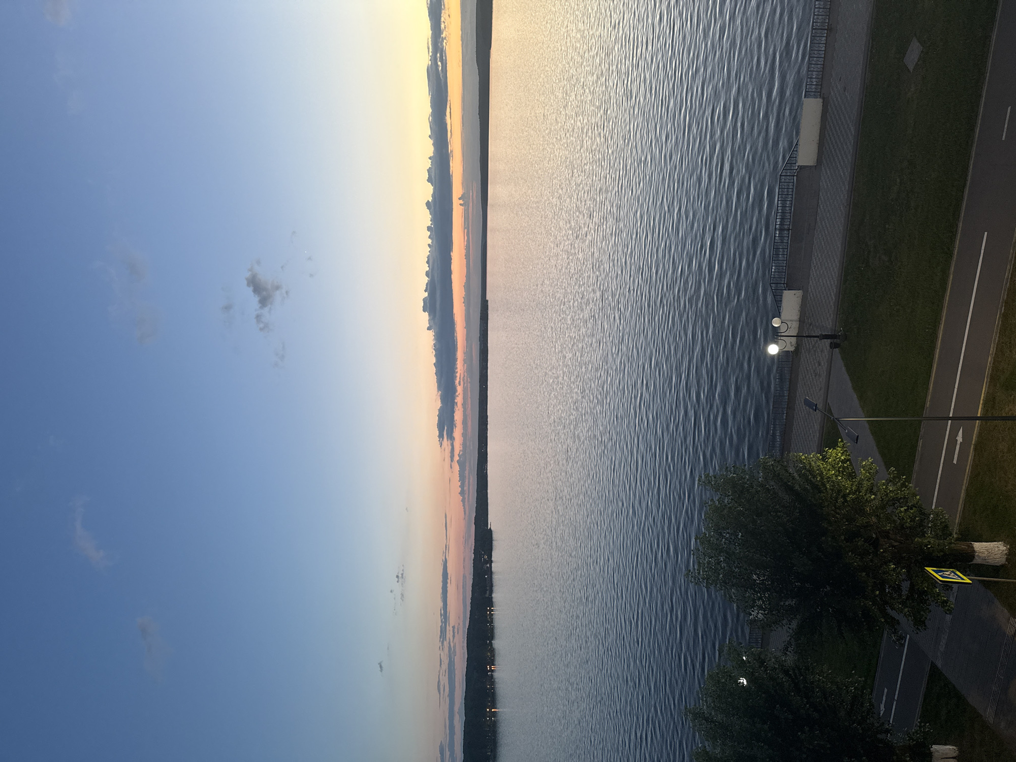 This image captures a serene twilight scene overlooking a wide body of water, likely the Votkinsk Reservoir, given the location context. The viewpoint is elevated, looking across the water towards a distant shoreline.
The sky occupies the left half of the frame, transitioning from a clear, deep blue overhead to soft hues of yellow, orange, and light blue closer to the horizon, indicative of either sunrise or sunset. A few small, scattered dark clouds are visible against the blue sky.
The large expanse of water, possibly a lake or wide river, fills the middle and right side of the image. Its surface is covered with subtle, regular ripples, reflecting the ambient light and appearing brighter on the side where the sun's low angle is most prominent.
Along the immediate right edge of the frame, a paved promenade or embankment runs parallel to the water's edge. A dark metal railing separates the walkway from the water. Further inland, a strip of green grass borders an asphalt road. The road features clear white markings, including a solid line, a dashed line, and directional arrows pointing away from the viewer.
Artificial lighting is present: a street lamp with two glowing globes illuminates the promenade, and another street lamp is partly visible further down the road, obscured by foliage. Several trees with dark green leaves are prominent in the foreground and mid-ground on the right, one with its lower trunk painted white. A yellow triangular sign with a black border and a black pictogram of a pedestrian crossing is visible near the road, partially hidden by a tree.
Across the distant horizon, a dark, narrow line of land or trees is visible, with faint specks of light suggesting distant settlements or structures. No people or specific activities are discernible, contributing to the tranquil atmosphere of the scene. The overall impression is one of peaceful dusk or dawn over a significant waterfront area.