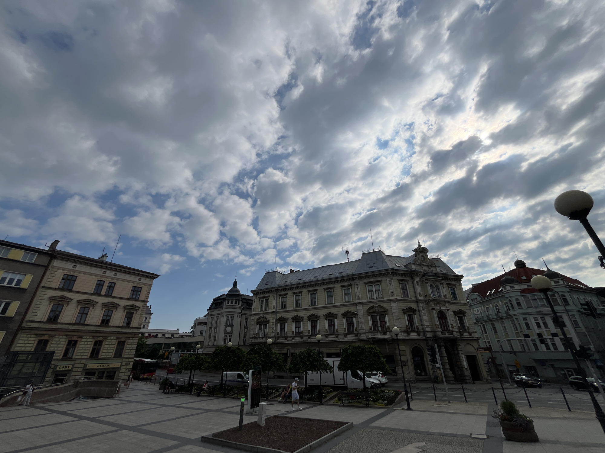 The image captures an outdoor scene in a European-style city square, likely during the daytime under a cloudy sky. The sky is filled with dramatic, textured clouds, with patches of sunlight breaking through, suggesting either early morning or late afternoon.
The foreground features a paved square with a few planters and lampposts. Several people are visible, appearing to be casually strolling or sitting on benches.
The background is dominated by ornate, historic buildings with distinct architectural styles, featuring multiple stories, large windows, and decorative facades. One building, in particular, stands out with its intricate detailing and mansard roof. Across the street, more buildings are visible, some with red-tiled roofs.
A few vehicles, including buses and cars, are seen on the street adjacent to the square, indicating an active urban environment. Some trees are planted along the square, adding a touch of greenery.
There is some visible text on the buildings, including "LODY" and "DESERT" on one of the storefronts, suggesting shops or cafes. The overall impression is one of a vibrant city center on a day with dynamic weather.