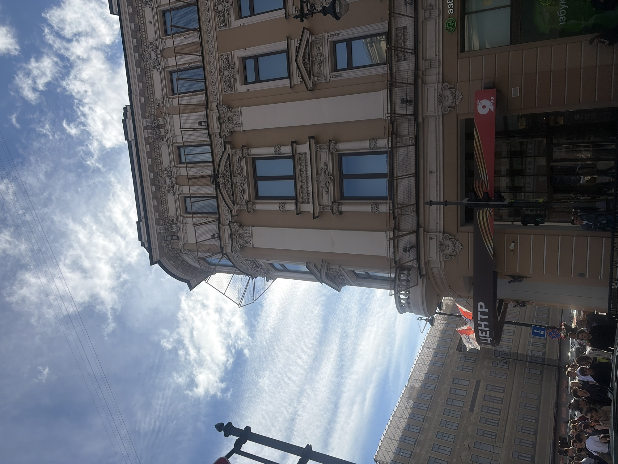 The image depicts a view of a street scene in Saint Petersburg, Russia, focusing on the facade of a multi-story building. The building is ornate, with classical architectural details, including decorative cornices, columns, and balconies. Several windows are visible, some reflecting the blue sky and clouds. A prominent sign with the word "ЦЕНТР" (Center) in Russian is displayed above an entrance. Red and white flags, possibly national flags, are flying from the building. To the left, another building with numerous windows is partially visible, appearing to be under construction or renovation, covered in netting. A street light and traffic signals are also present. In the foreground, a crowd of people is gathered on the sidewalk, some looking towards the buildings, others appearing to be walking or conversing. The sky is a bright blue with wispy clouds. The overall impression is of a sunny day in a city with grand architecture.